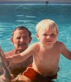 A man and a young boy in a swimming pool, smiling. The man has dark hair and a beard, the boy has blonde hair and is shirtless.
