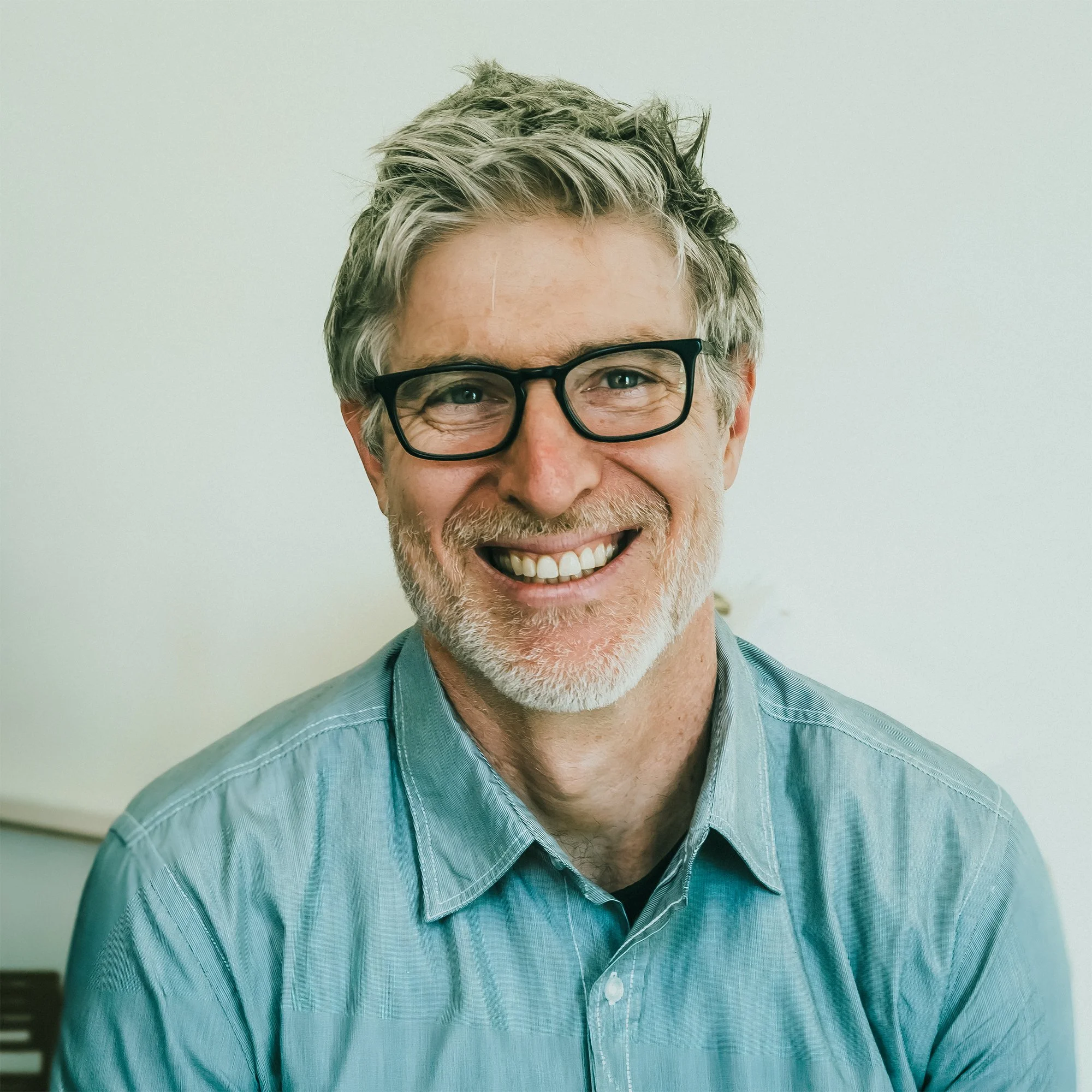 A smiling man with glasses, gray hair, and a beard wearing a light blue, slightly wet dress shirt standing against a plain, light-colored wall.