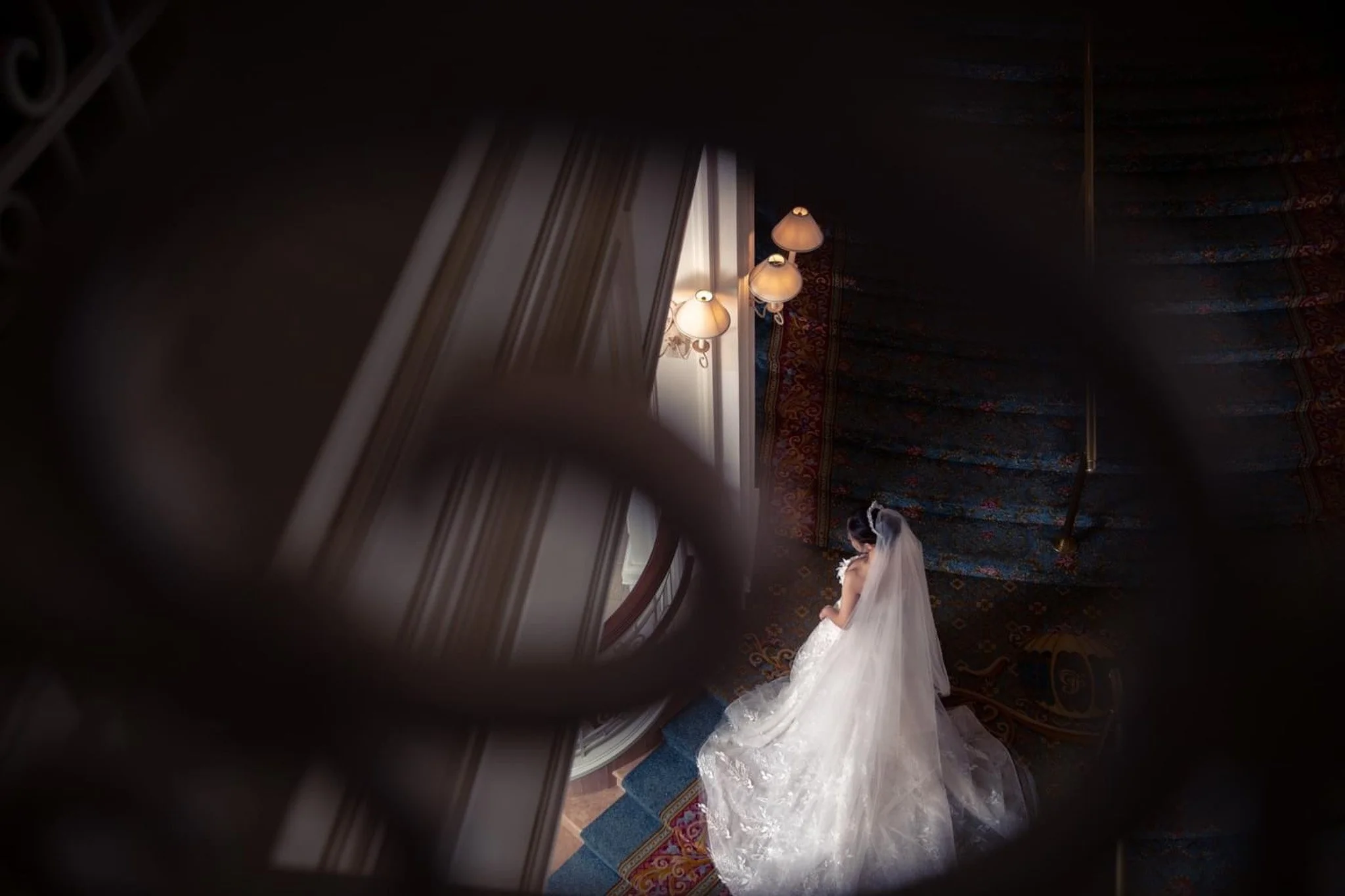 Bride in a white wedding gown and veil standing on a staircase, viewed from above through a decorative wrought iron railing.