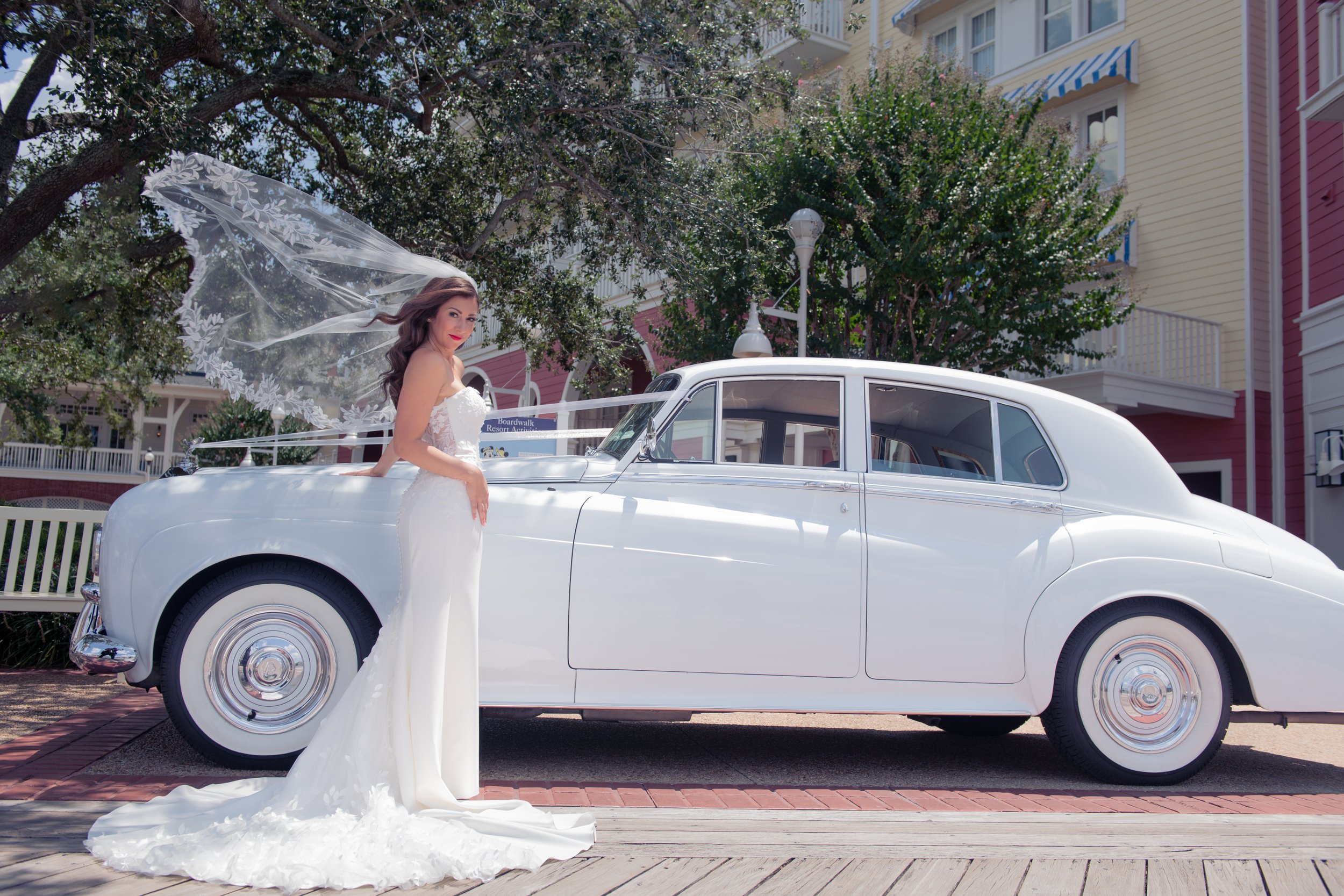 Bride in a white wedding dress with a long train standing beside a vintage white car on a city street.