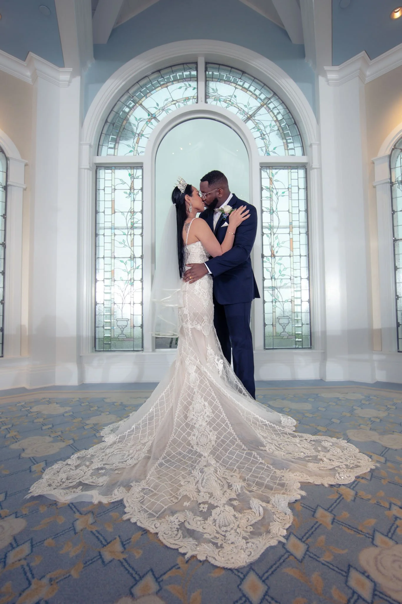 A bride and groom sharing a kiss in front of a large stained glass window in Disney's Wedding Pavilion, with the bride wearing a lace wedding gown with a long train and the groom in a navy suit.