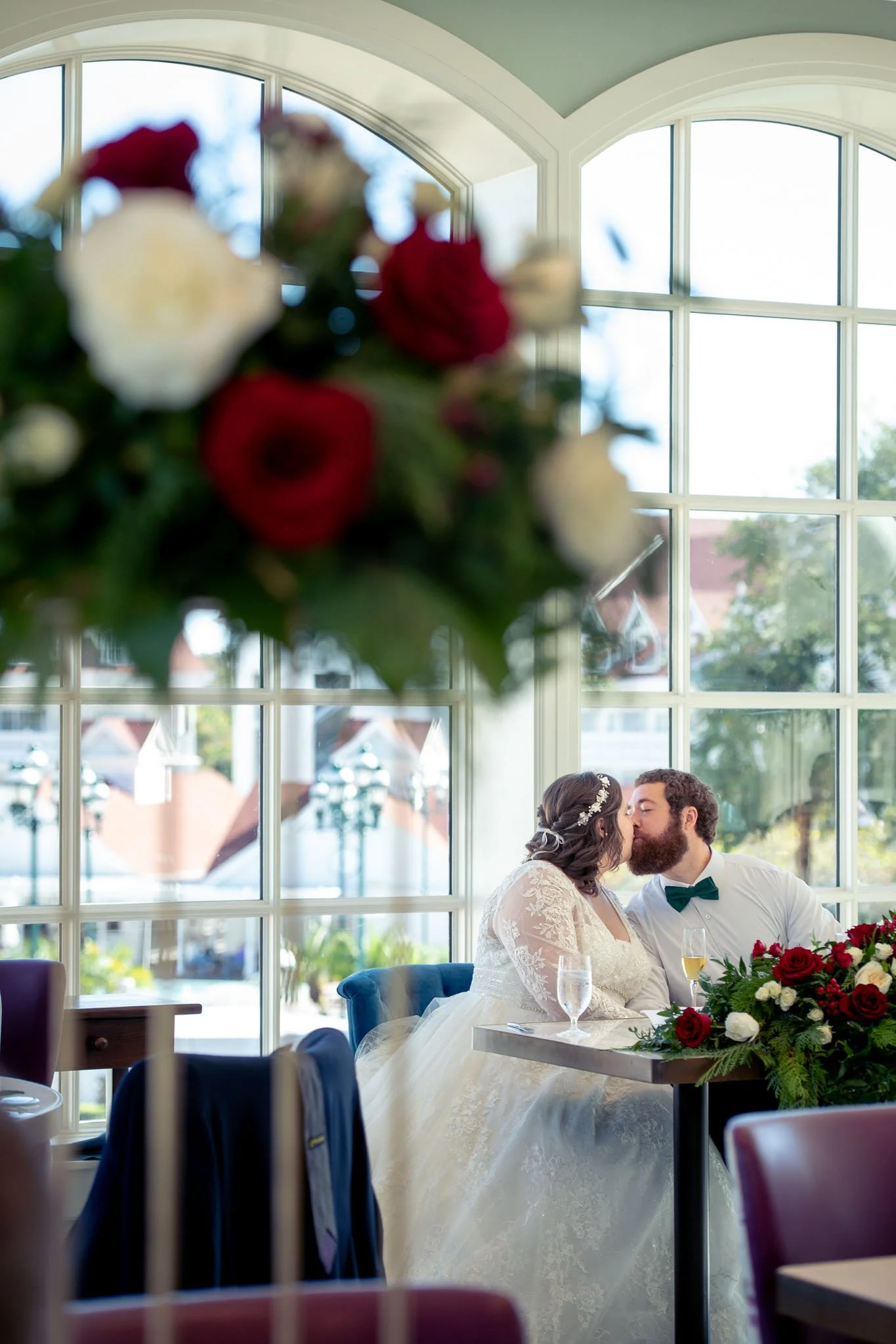 A bride and groom sharing a kiss at their wedding reception, sitting at a table with floral arrangements, with a large window in the background at Citricos at the Grand Flordian.