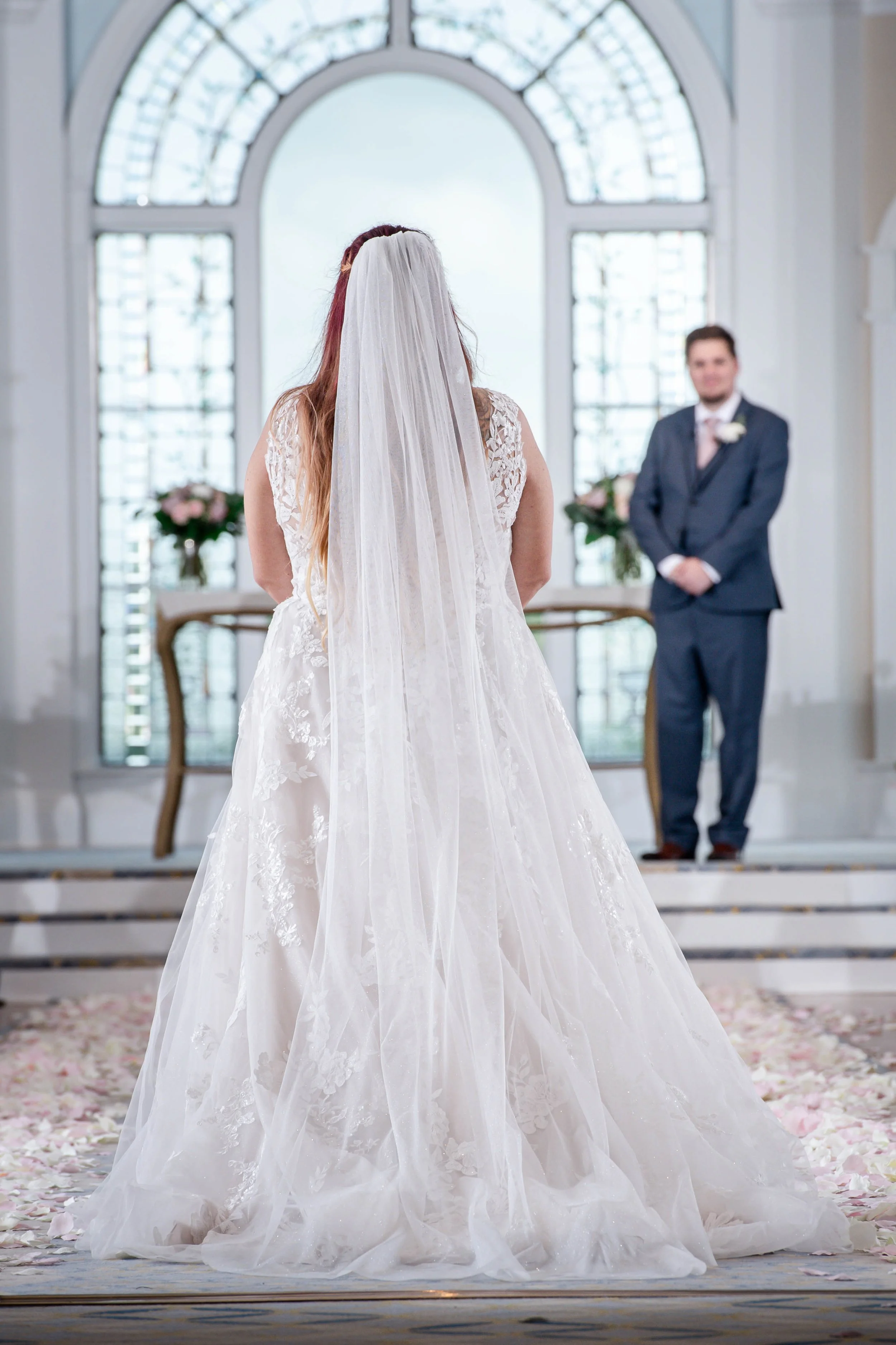 Bride in a wedding gown with a long veil facing groom in a formal suit at the altar during a wedding ceremony in a bright, elegant church with large arched windows, floral arrangements, and petals on the floor.
