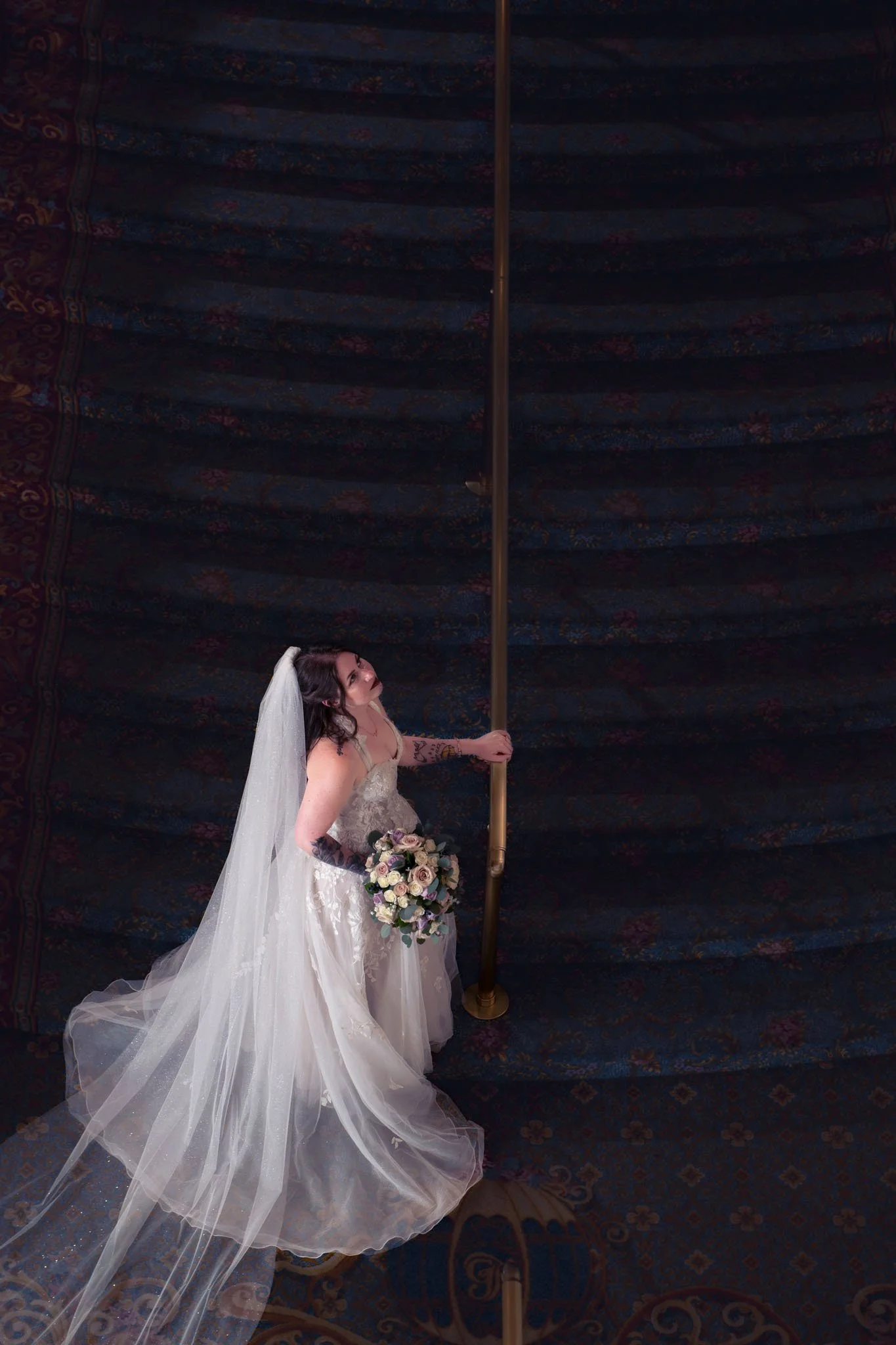 A bride in a wedding gown with a long train and veil, holding a bouquet, standing beside a pole on a patterned dark background.