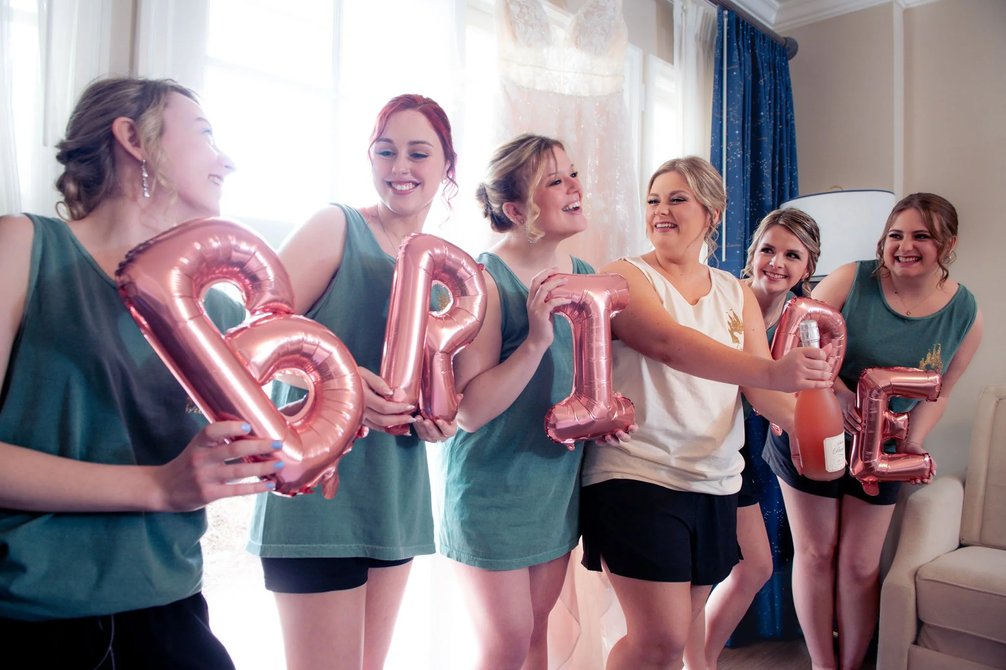 Group of women celebrating, holding pink balloons spelling LOVE, some holding a bottle of champagne, smiling and having fun indoors.