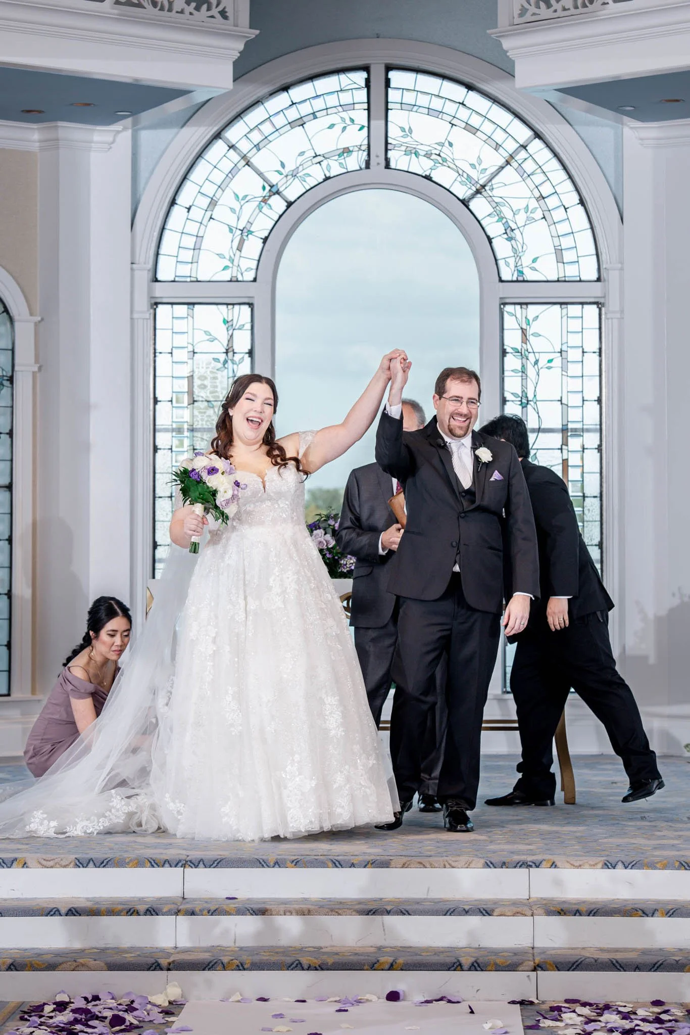 A newlywed couple holding hands and smiling on at Disney's Wedding Pavilion with a woman kneeling and adjusting the bride's dress, and other individuals in the background.