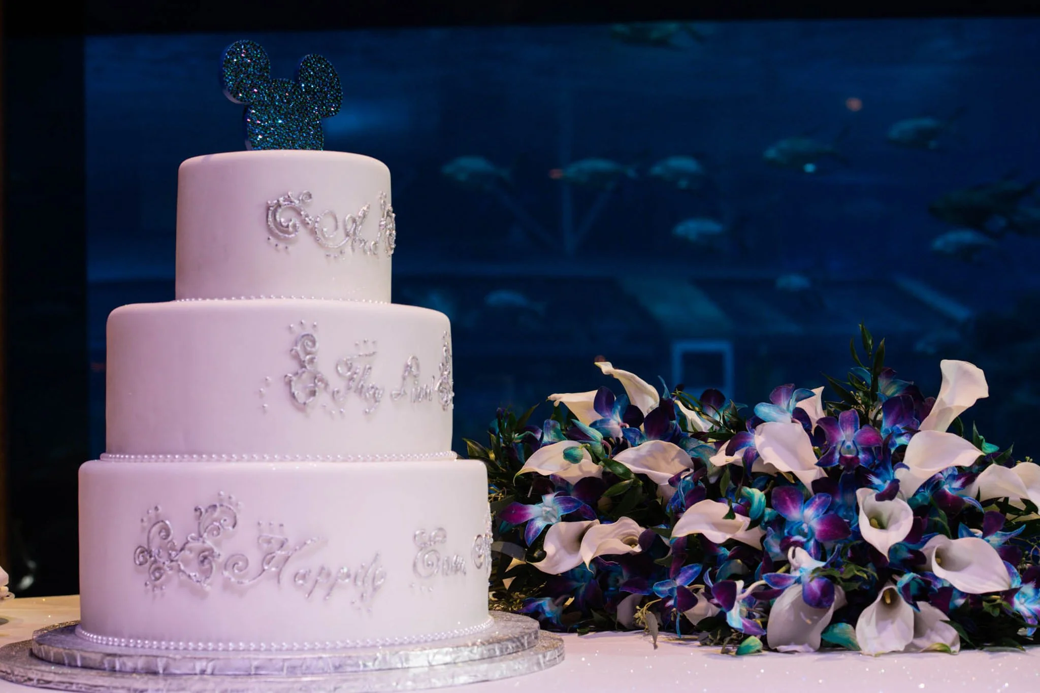 A three-tier white wedding cake with 'Love' and 'Happy' written on it, decorated with silver accents, topped with a glittery Mickey Mouse-shaped decoration, beside a large bouquet of white and purple calla lilies, with an aquarium tank in the backgro