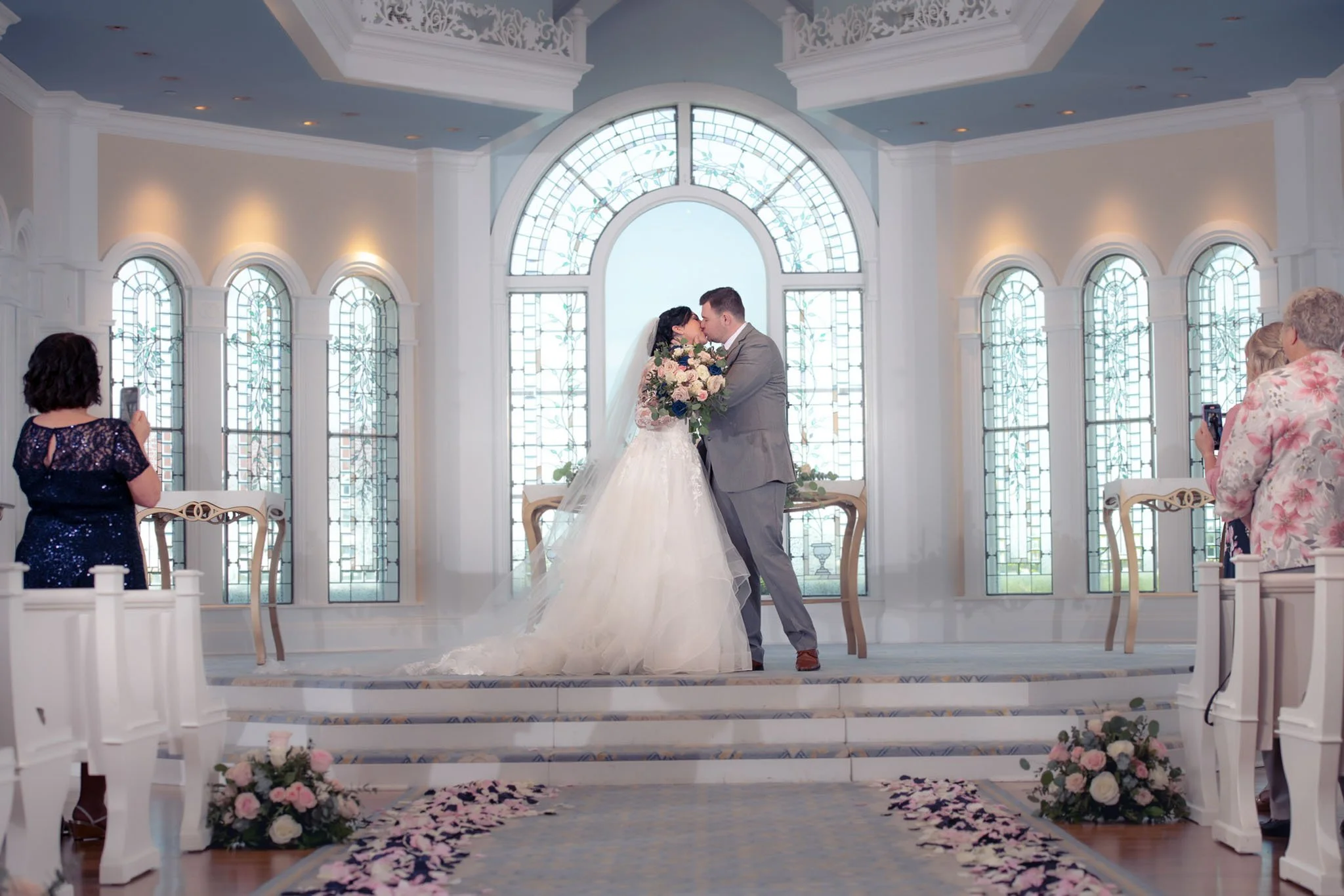 A bride and groom share a kiss at their wedding altar inside Disney's Wedding Pavilion with large stained glass windows. Their guests are taking photos, and floral arrangements decorate the steps.