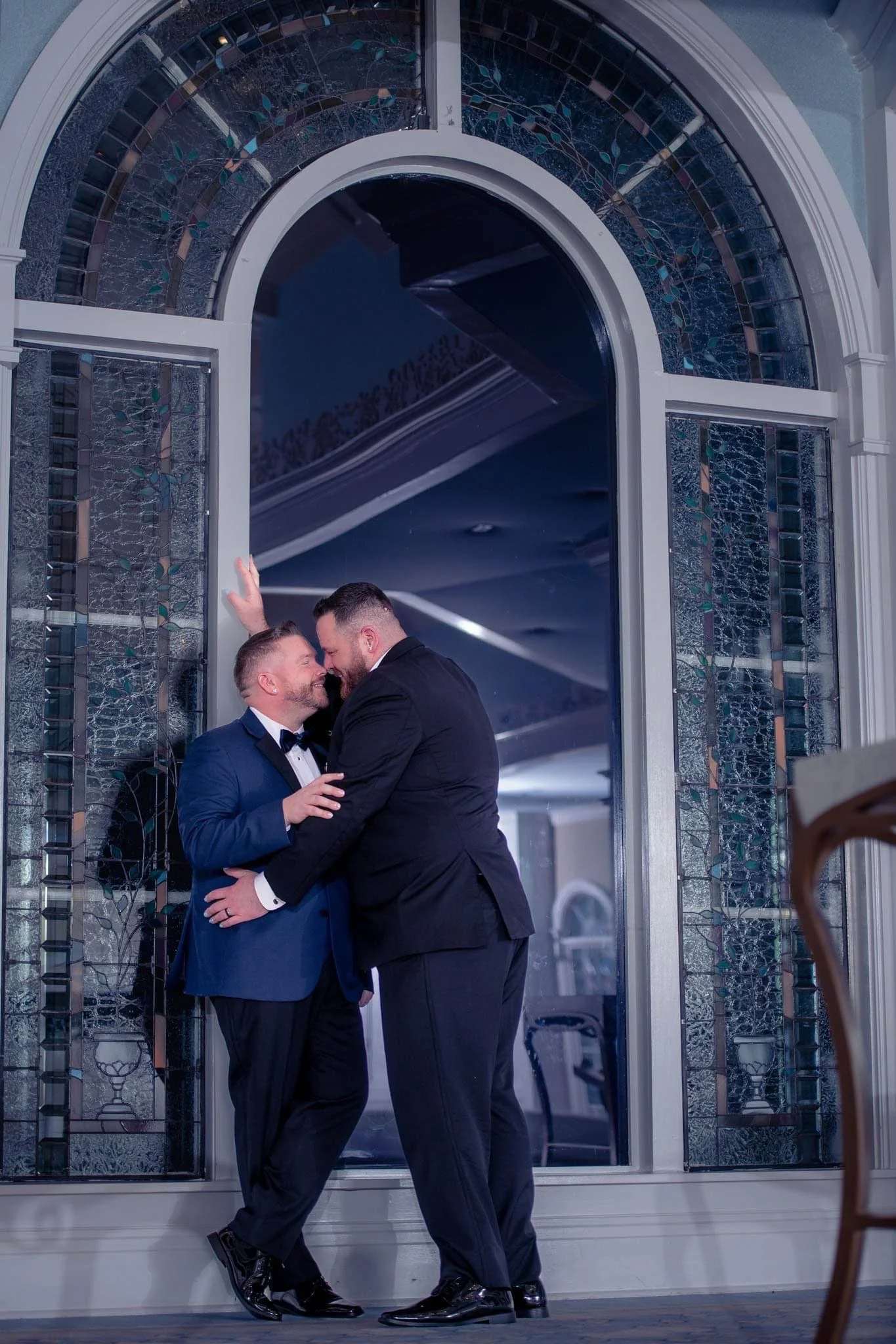 Two men in tuxedos sharing an intimate dance near a large stained glass window.