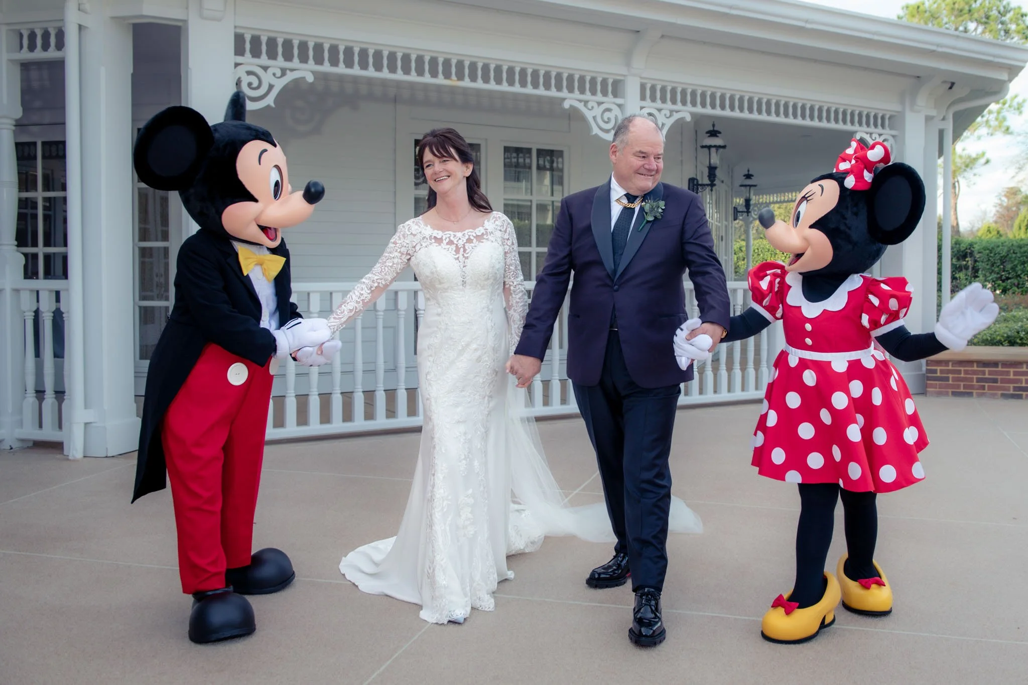 Bride and groom holding hands and smiling, flanked by Mickey and Minnie Mouse characters, in front of the White Hall at The Grand Floridian with a porch.