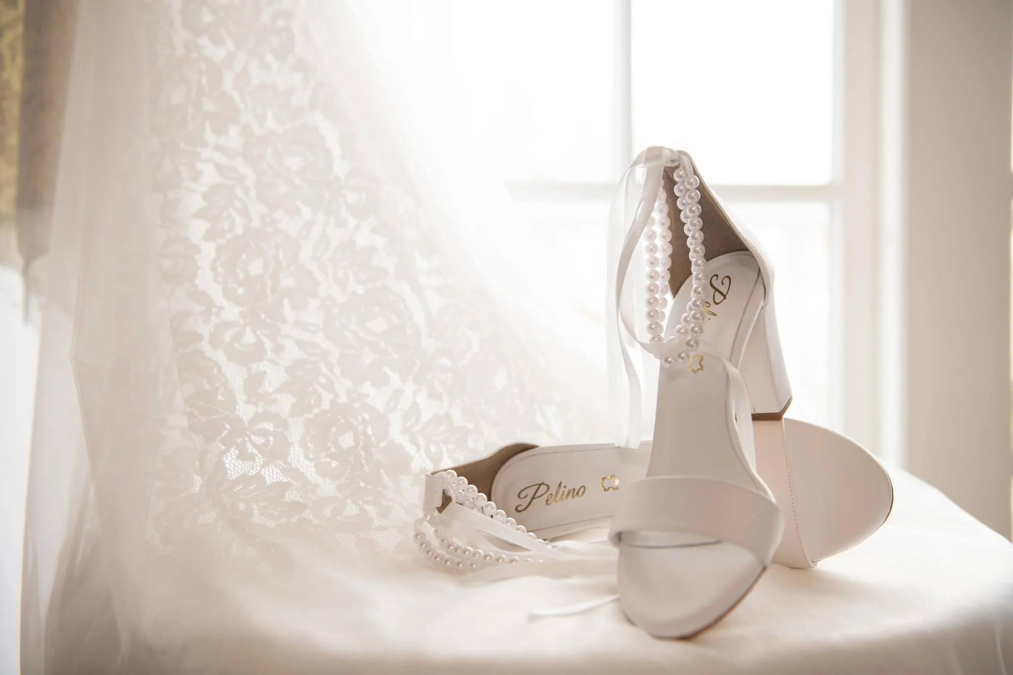 White wedding shoes with pearls and a pearl necklace on a white cushioned surface near a window.