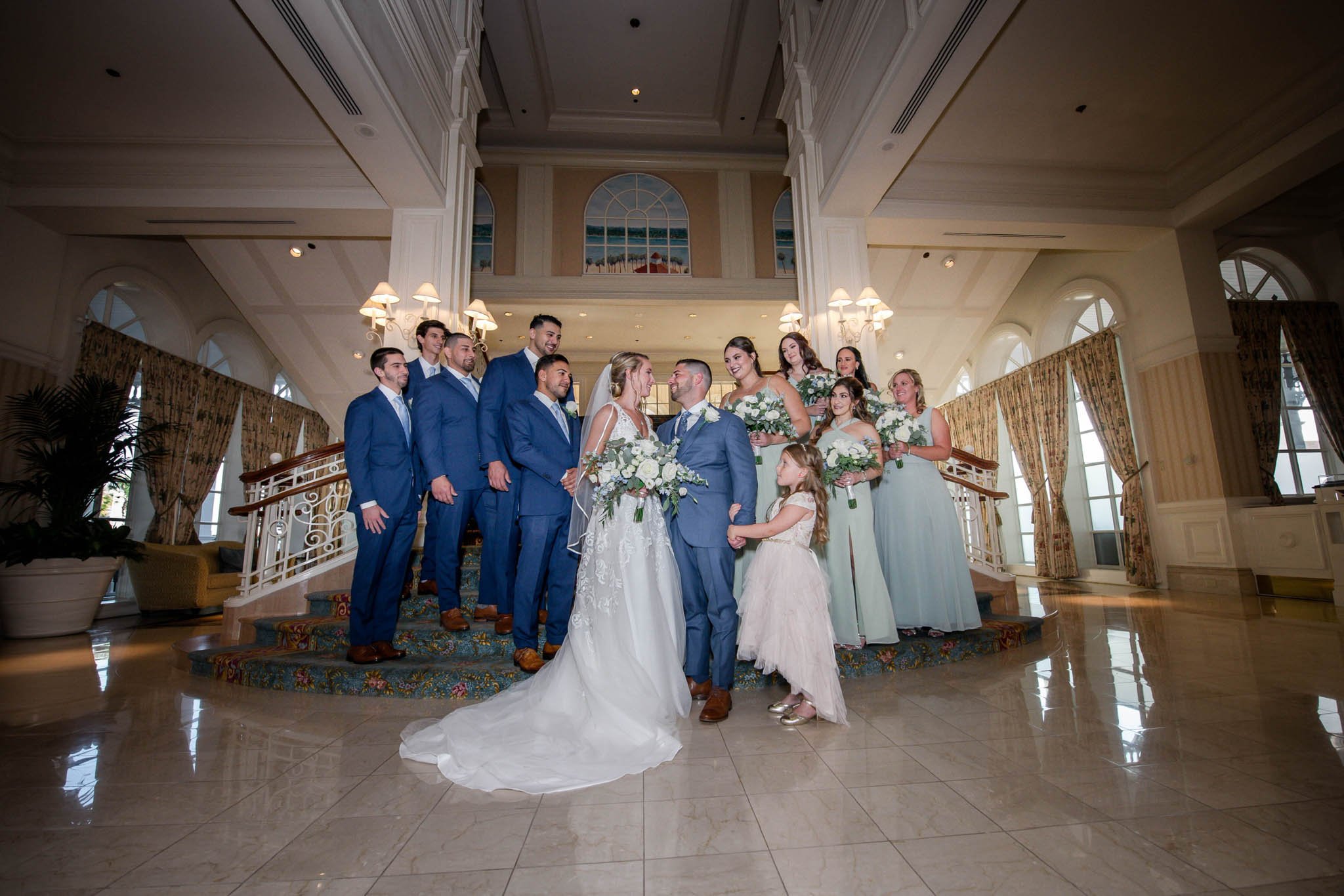 A wedding party with the bride and groom at the center, holding hands and surrounded by bridesmaids and groomsmen in a grand, elegantly decorated indoor venue with large windows, chandeliers, and curtains.