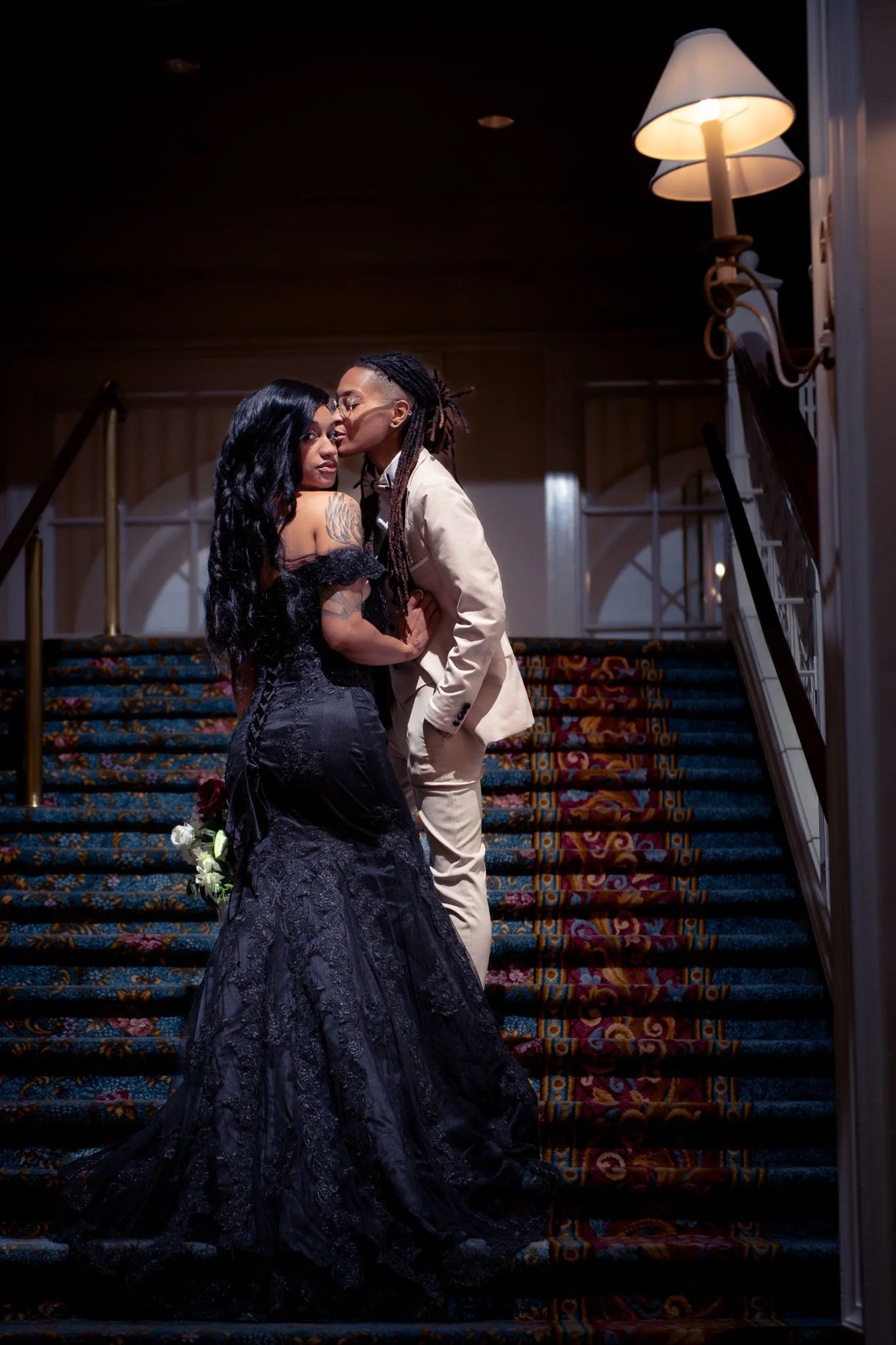 A couple stands on a staircase, the person in tuxedo kissing the woman in a black wedding dress on her cheek in an elegant indoor setting.