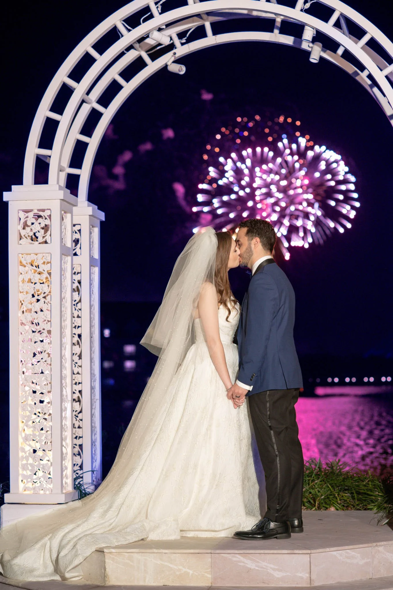 A bride and groom sharing a kiss under Disney's Picture Point archway with Magic Kingdom fireworks in the night sky behind them during their wedding celebration by the water.