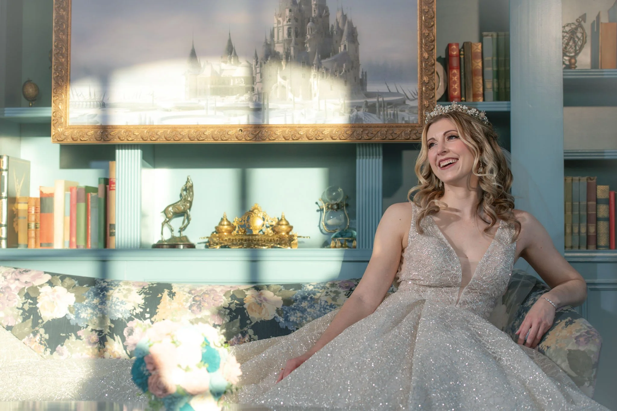 A woman wearing a sparkly gown and tiara, sitting on a floral sofa in a the Enchanted Rose with books and decorative items, smiling happily.