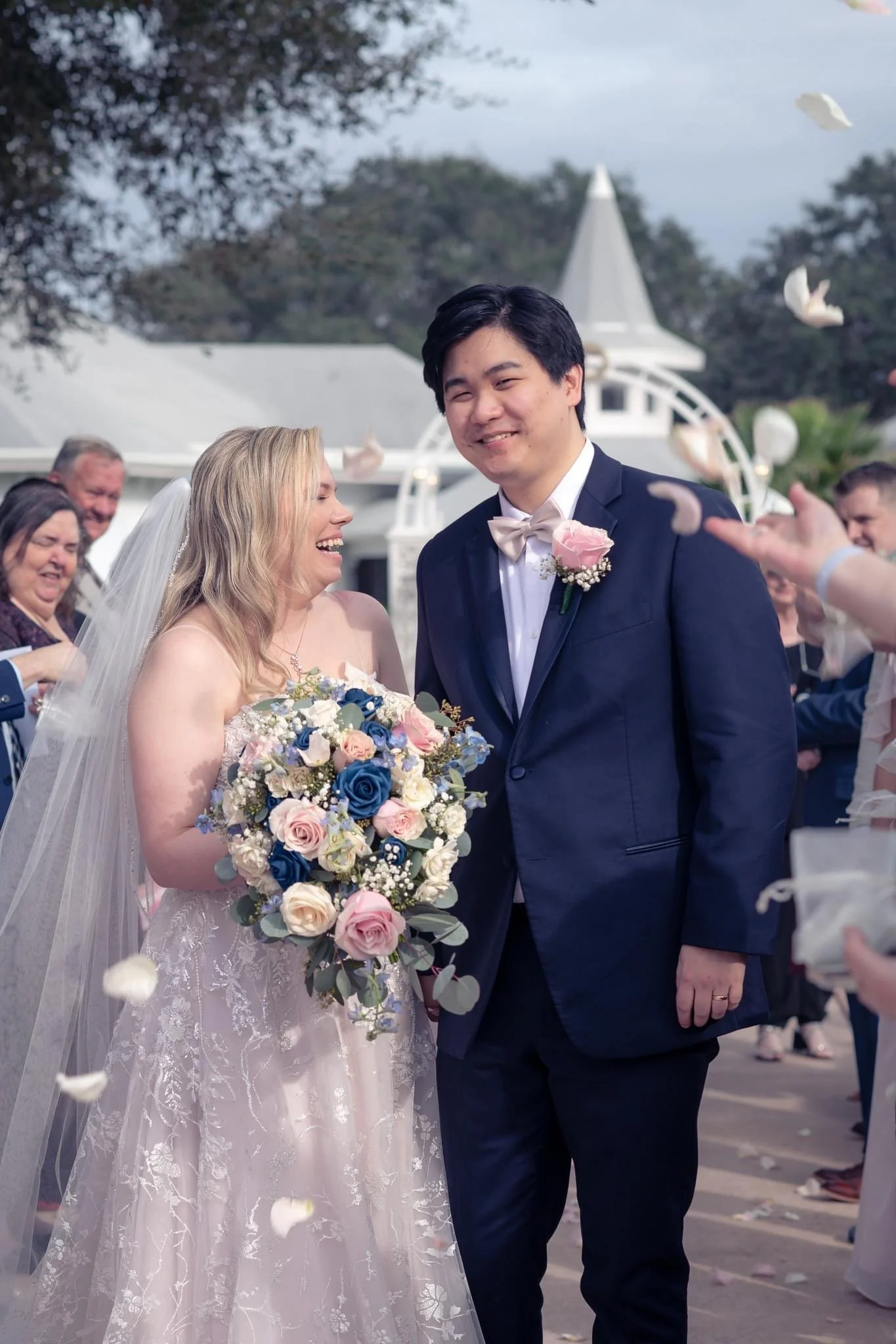 A newlywed couple sharing a joyful moment outdoors, surrounded by friends and family, with floral petals in the air during a wedding celebration at Disney's Wedding Pavilion.