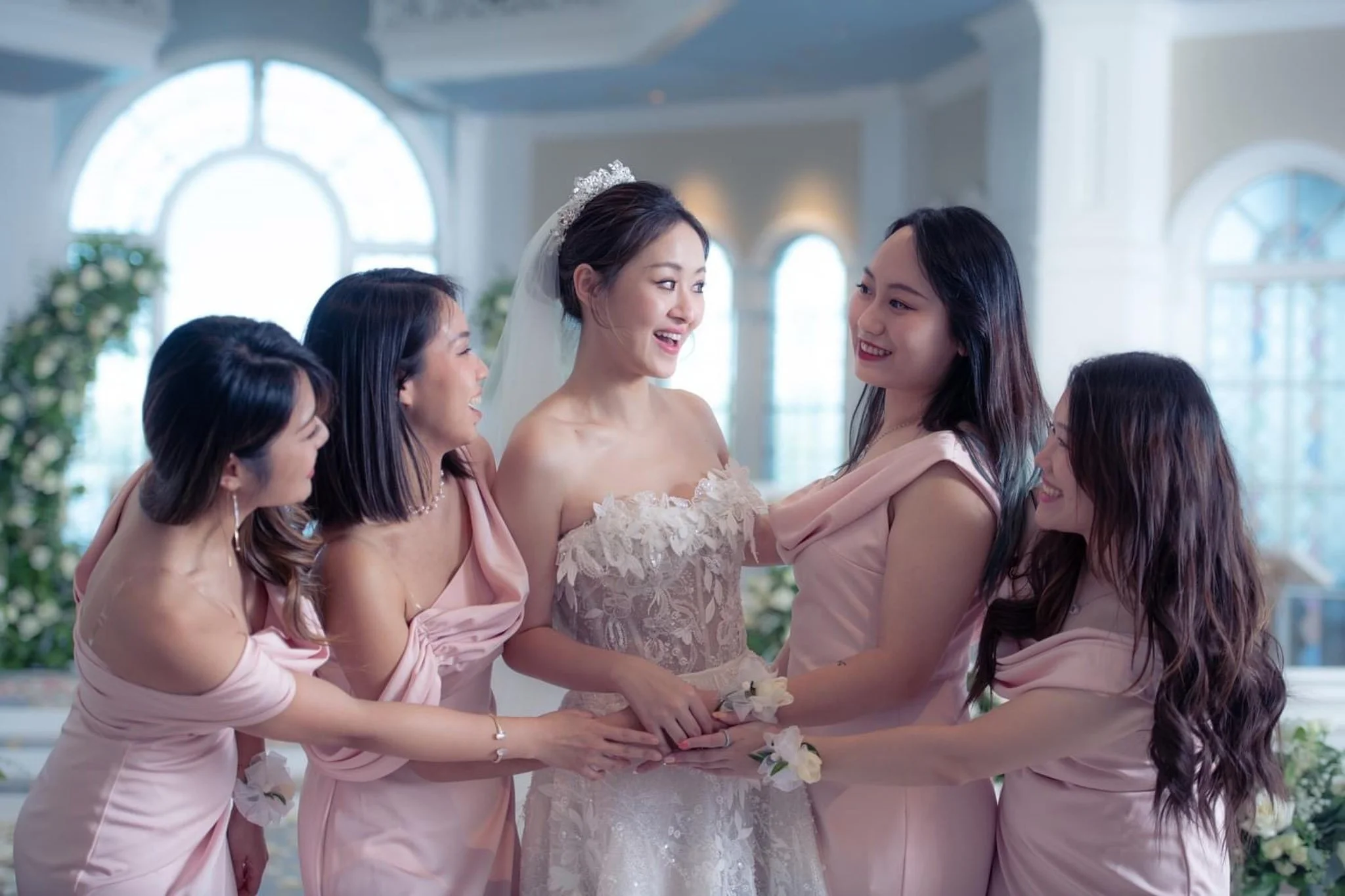 A bride in a wedding dress surrounded by five bridesmaids in pink dresses, all smiling and holding hands in a bright, at Disney's Wedding Pavilion with large arched windows.