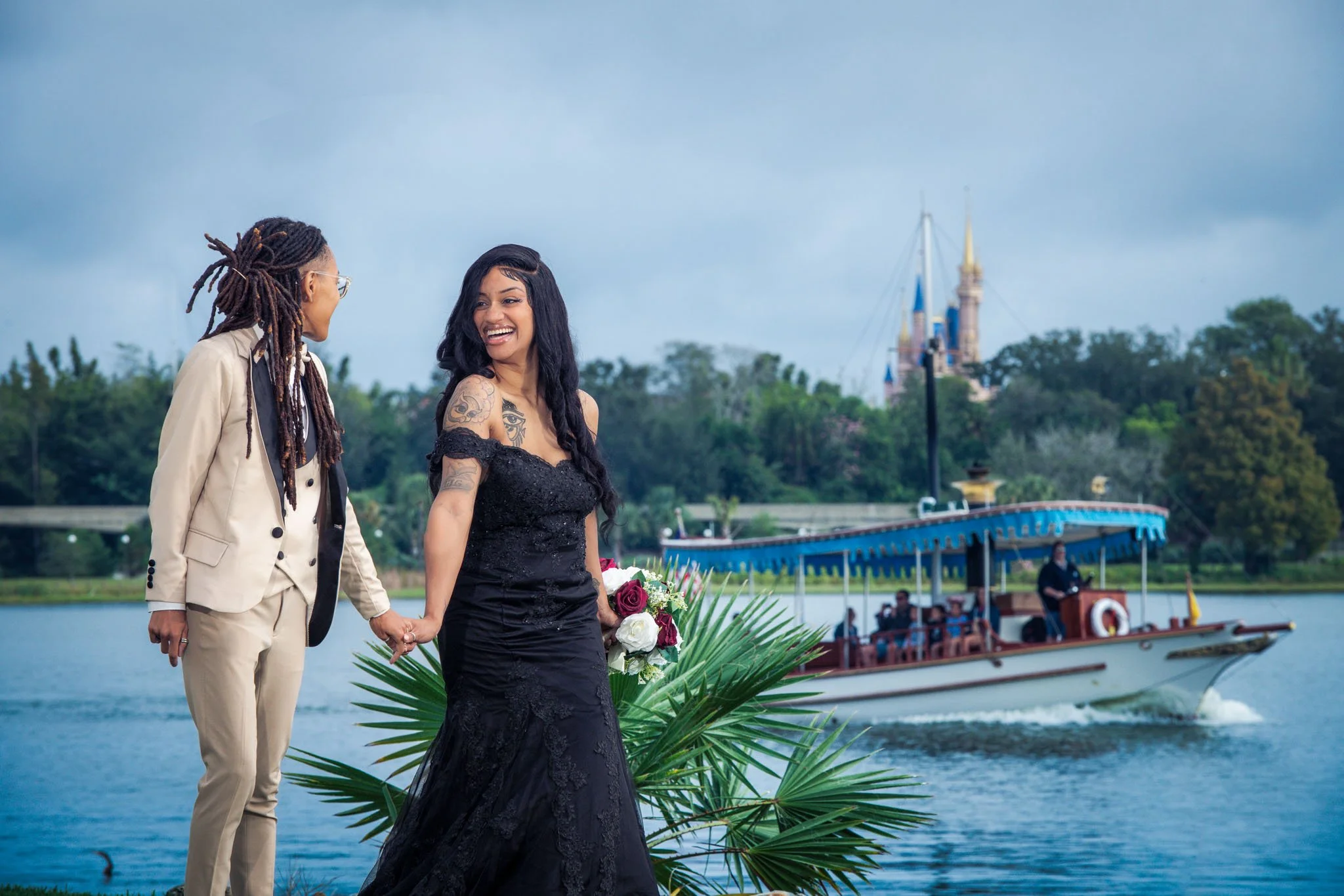 Happy couple at the Grand Floridian with Cinderella's castle by the water, with a boat and lush green trees in the background.