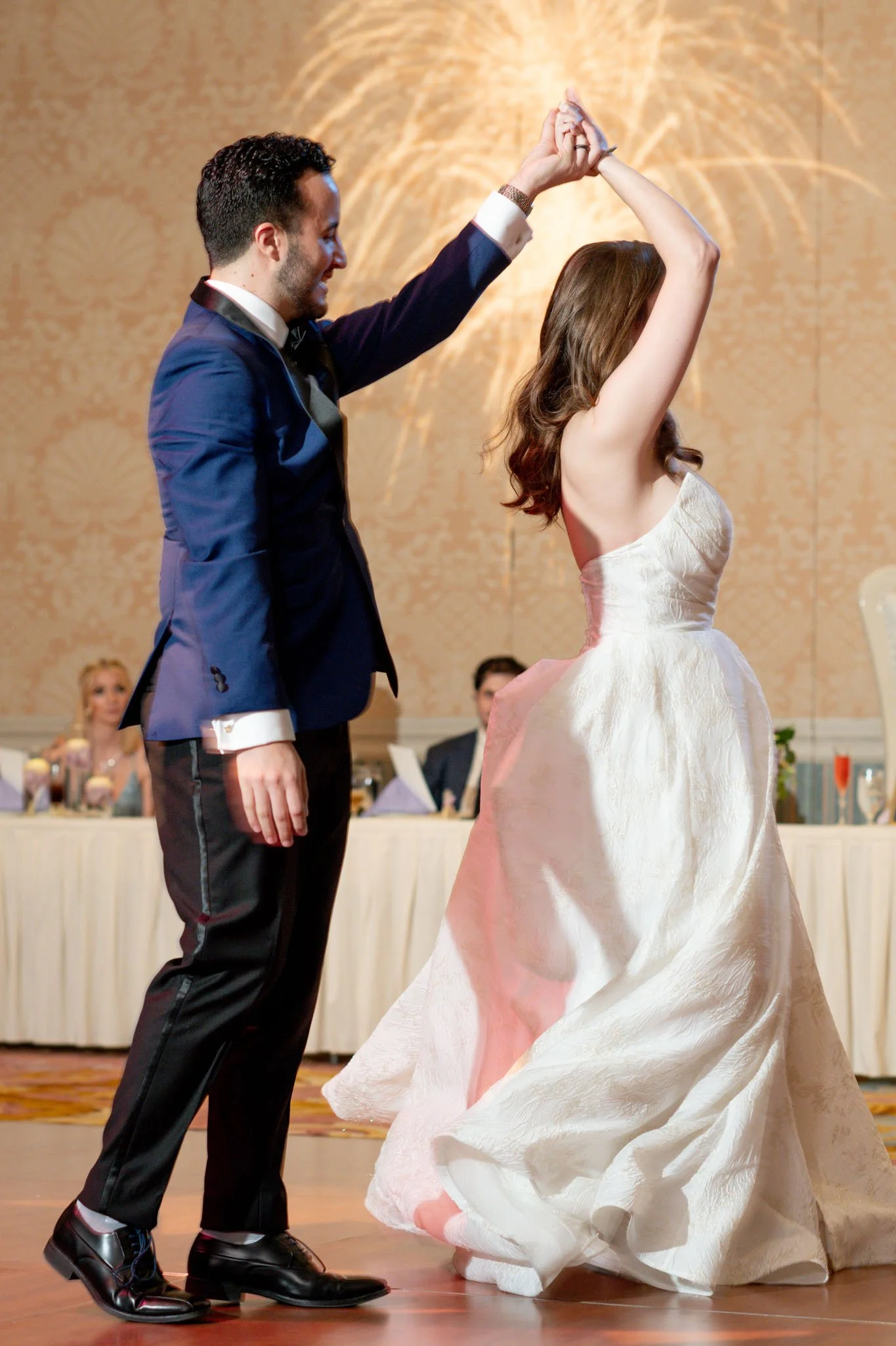 A couple dancing at their wedding reception, with the groom in a blue suit and the bride in a white strapless gown, with other guests seated in the background.