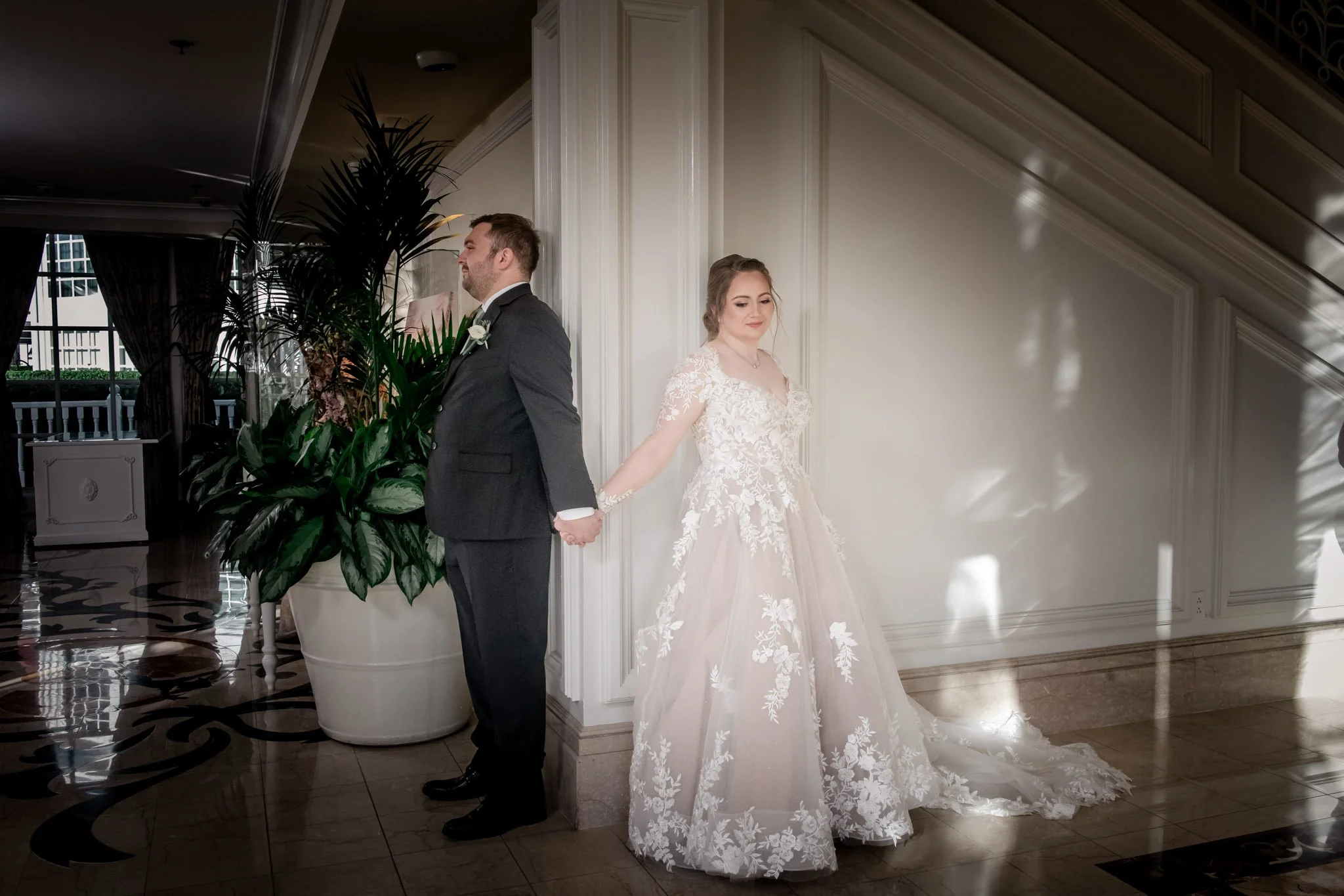 Bride and groom stand back to back, holding hands, in an elegant indoor setting with white paneled walls and sunlight reflections.