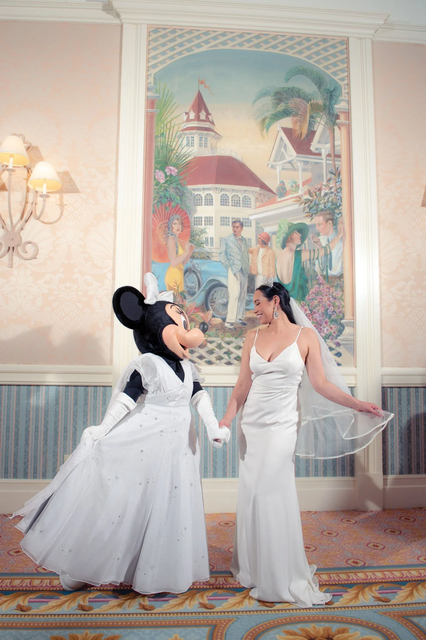 A woman in a white wedding dress and veil holding hands with a person dressed in a Mickey Mouse costume, also in a white dress, in an elegant room with a large colorful mural at the Grand Floridian on the wall behind them.