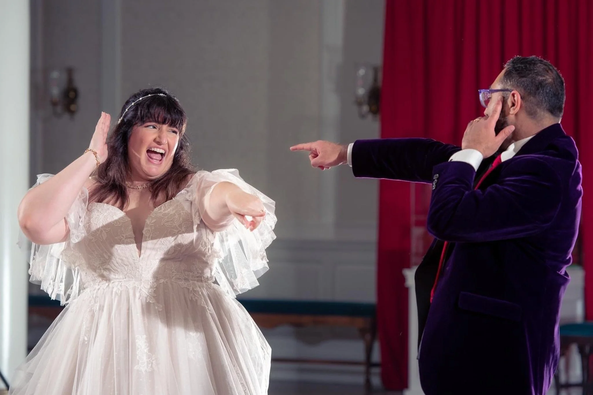 A woman in a wedding dress laughing and covering her ears while a man in a suit points and appears to be joking or teasing her.