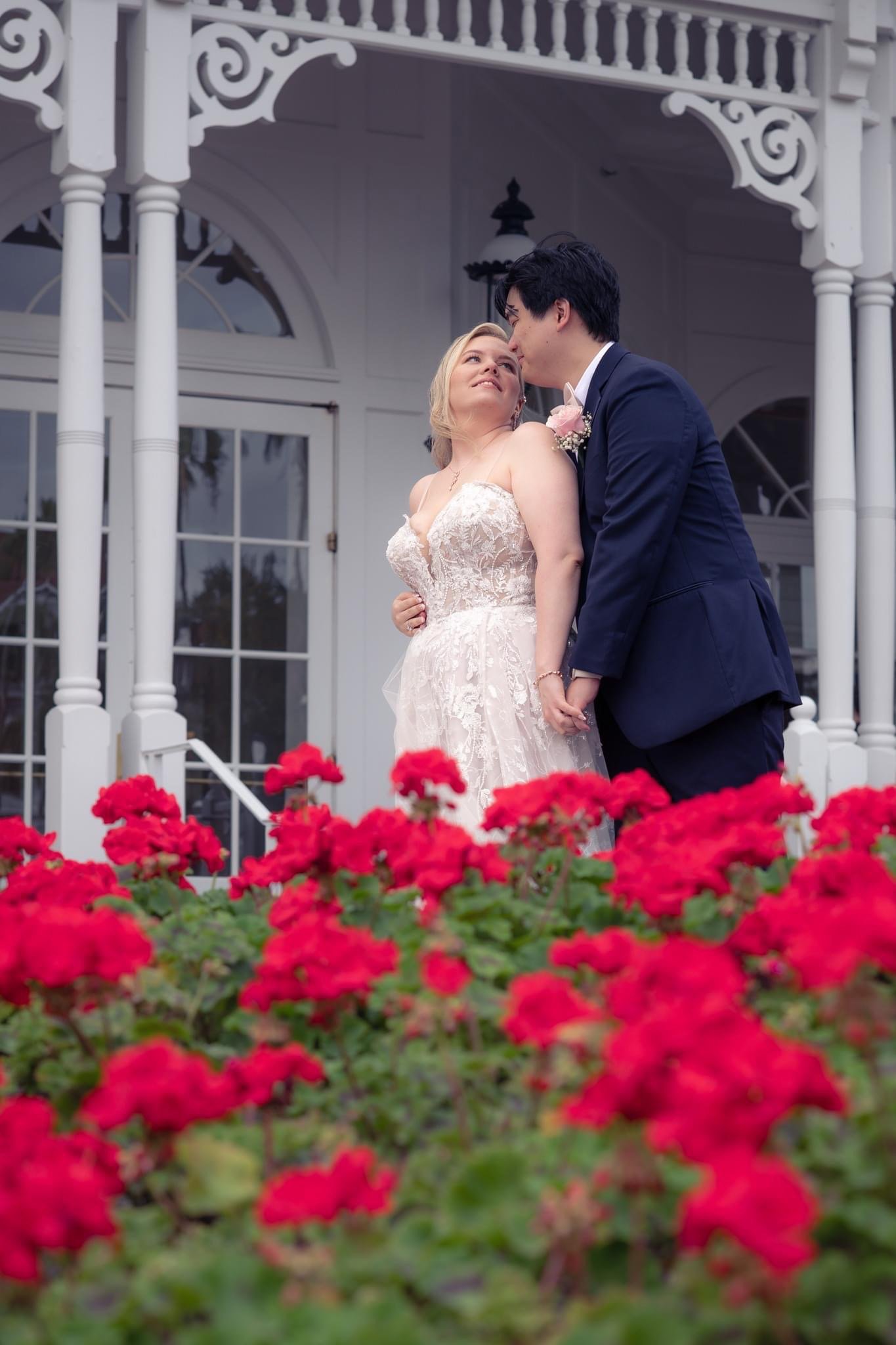 A newlywed couple sharing a romantic moment on the porch of a white, ornate house surrounded by bright red flowers at The Grand Floridian.