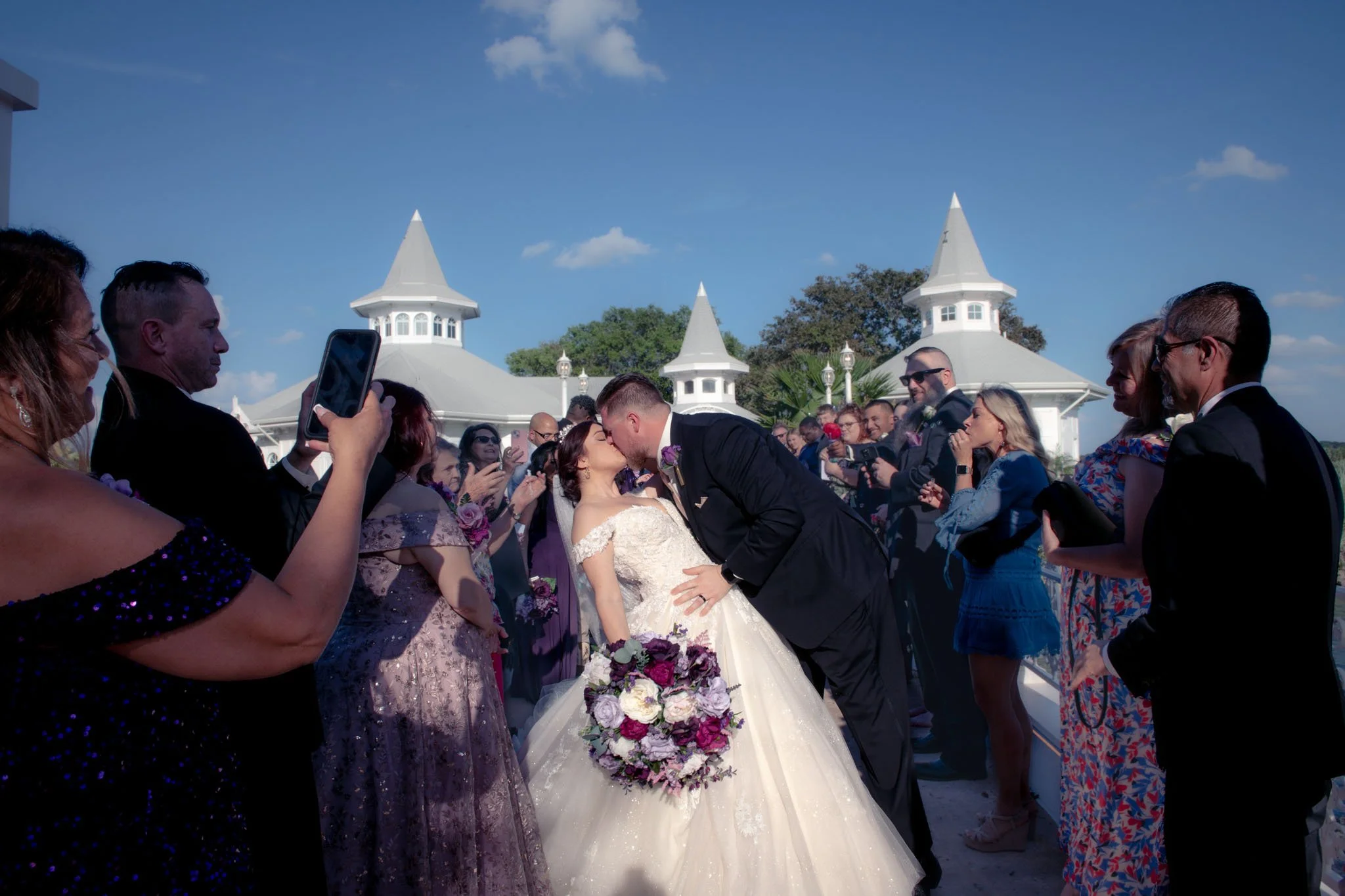A bride and groom kiss during their  wedding ceremony with guests around them taking photos and videos. The bride holds a large bouquet of purple, white, and pink flowers. The wedding is set against a background of white gazebos and a blue sky.