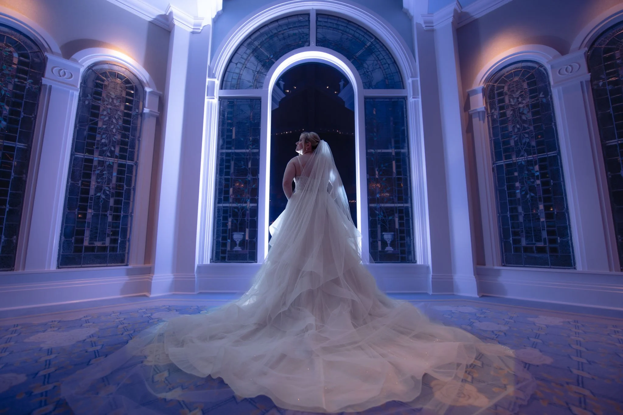 Bride in a wedding dress and veil standing in front of large stained glass windows in a brightly lit, elegant interior with white walls and blue accent lighting at Disney's Wedding Pavilion.