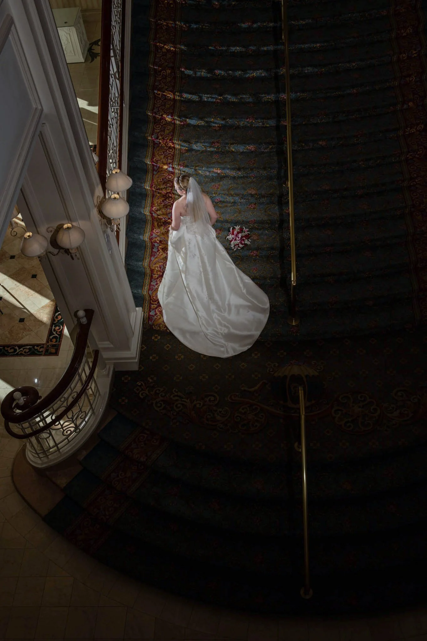 A bride in a white wedding gown walking down a grand staircase on a dark patterned carpet, holding a bouquet of flowers.