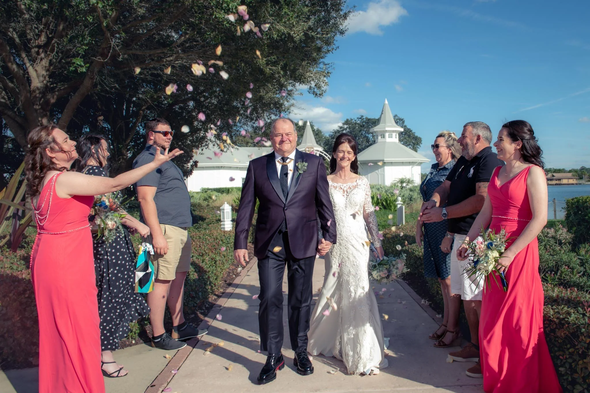 A bride and groom walking down a garden path at their outdoor wedding, surrounded by friends and family throwing flower petals, with a bright blue sky and a white gazebo in the background.