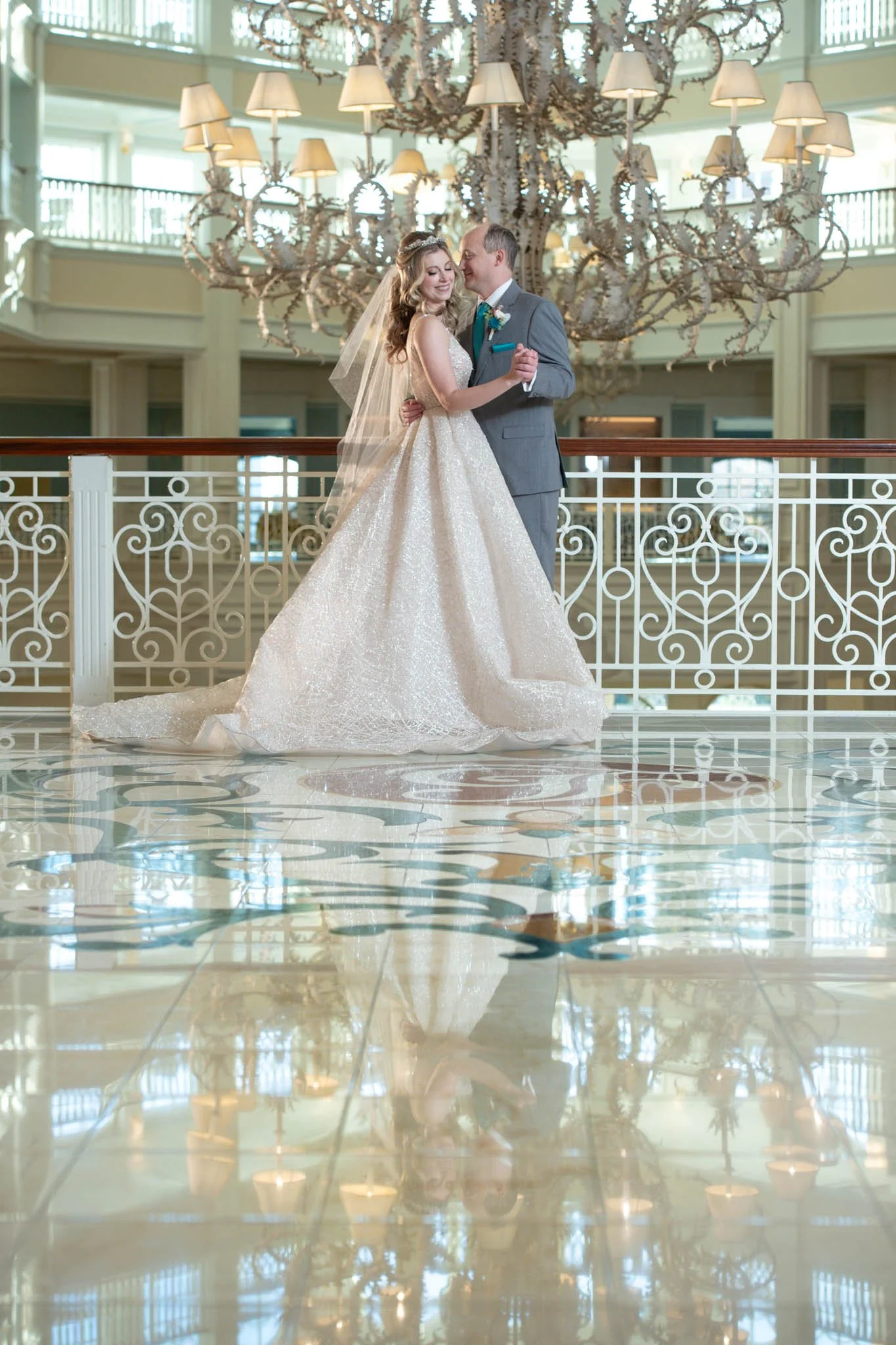 A bride and groom dancing together in a grand, well-lit hotel lobby with a large chandelier overhead and a decorative railing behind them.