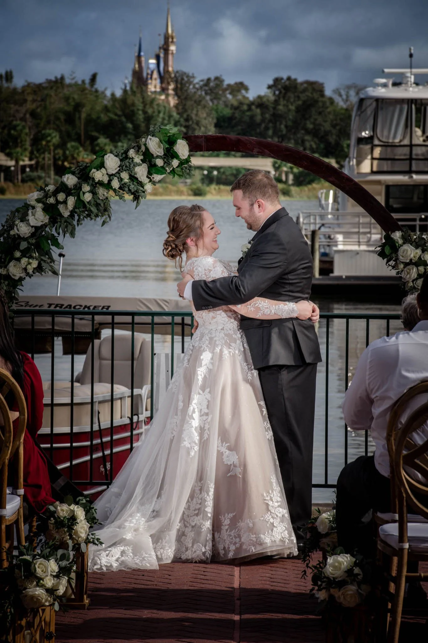 A bride and groom are sharing a dance or moment during their outdoor wedding ceremony by a river, with a castle in the background. The bride is wearing a white lace wedding gown, and the groom is in a black suit. They are under a floral archway decor