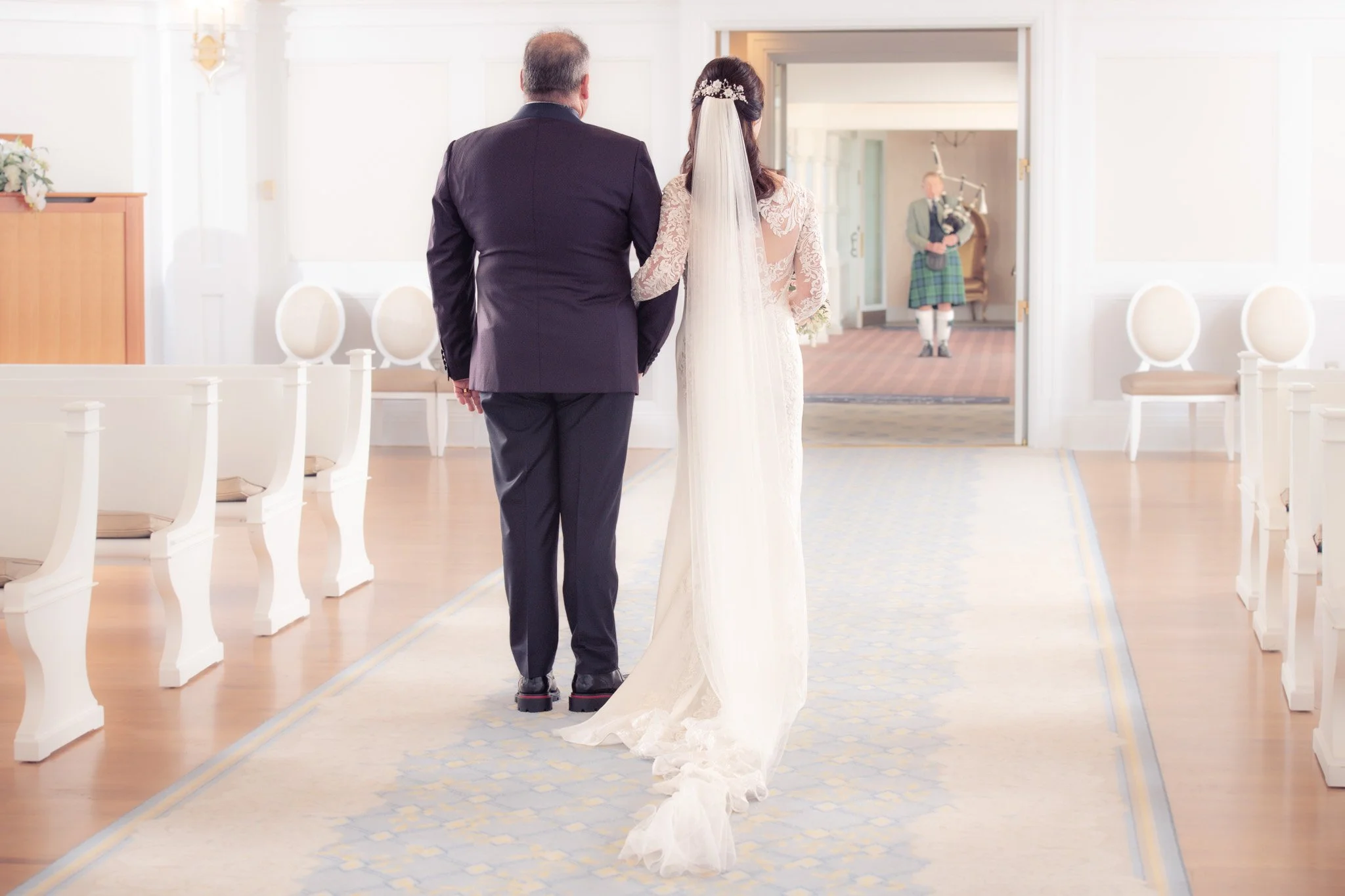 Bride and man walking down an aisle in a wedding ceremony in a bright, elegant room with white walls and wooden floors, while a woman in traditional Scottish attire plays bagpipes in the background.