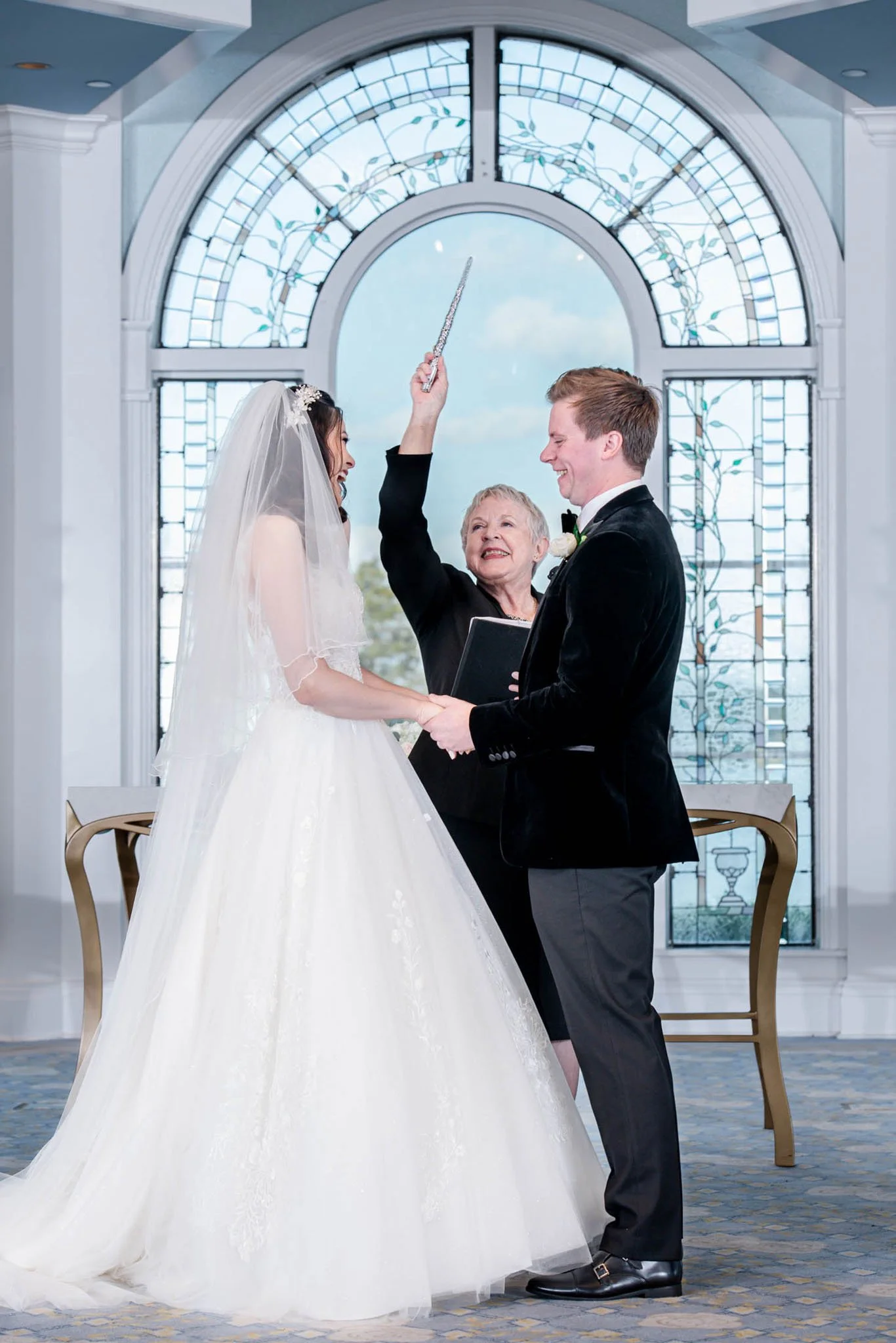 A bride and groom exchange vows during their wedding ceremony, with a officiant, alovelyceremony, holding a wand above them, in front of a large stained-glass window.