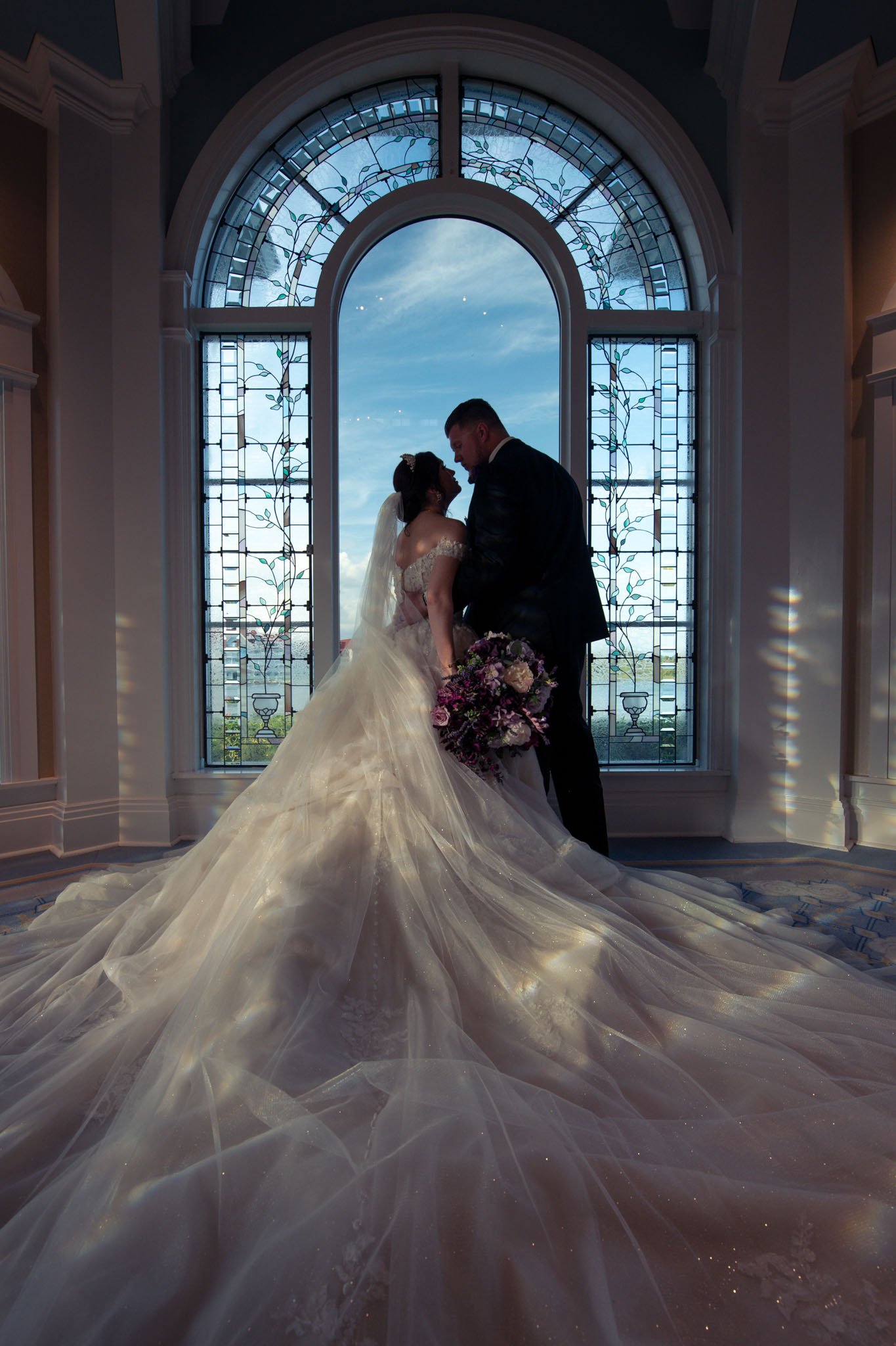 A bride in a white wedding gown and a groom in a black suit stand close together in front of a large arched window with stained glass. The bride holds a bouquet of flowers and the scene is backlit by natural light, creating a silhouette of the couple