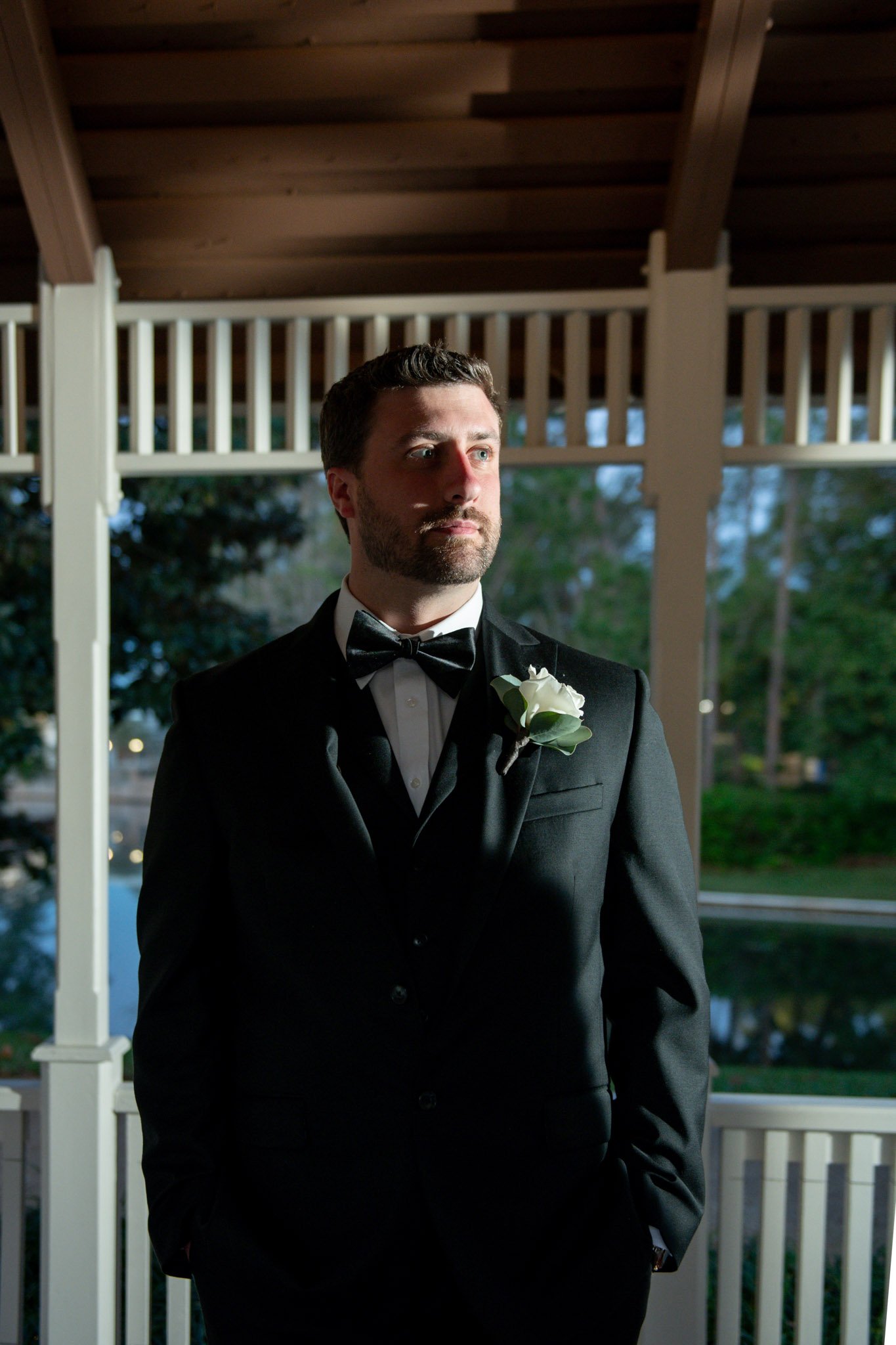 A man dressed in a black tuxedo with a bow tie and a white flower boutonniere standing on a porch during the evening with trees and water in the background.