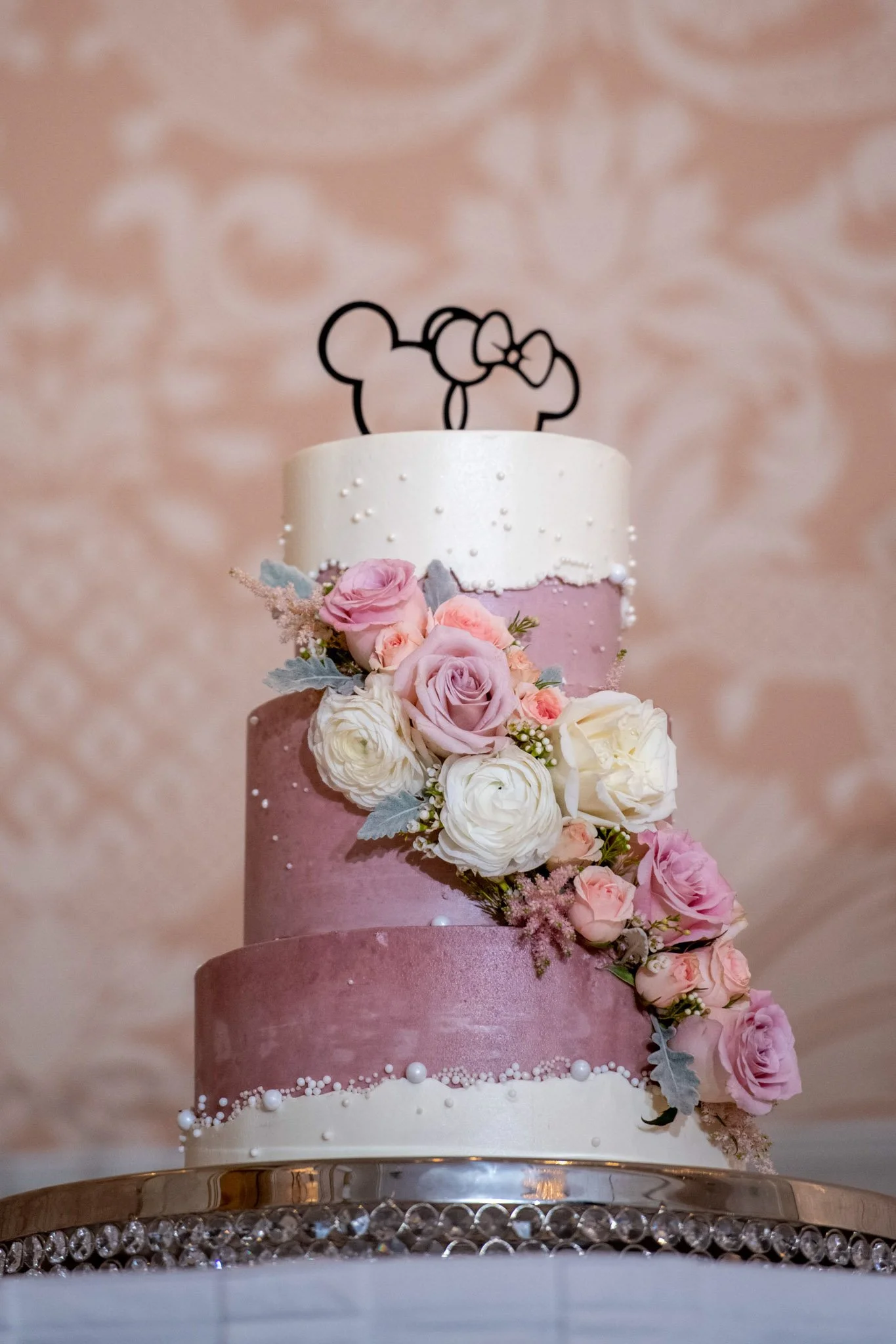 Three-tier wedding cake with pink and white icing, decorated with fresh pink and white roses, other small flowers, and grey leaves. A black outline of Mickey Mouse with a bow on his head is on top of the cake. The cake sits on a decorative silver tra
