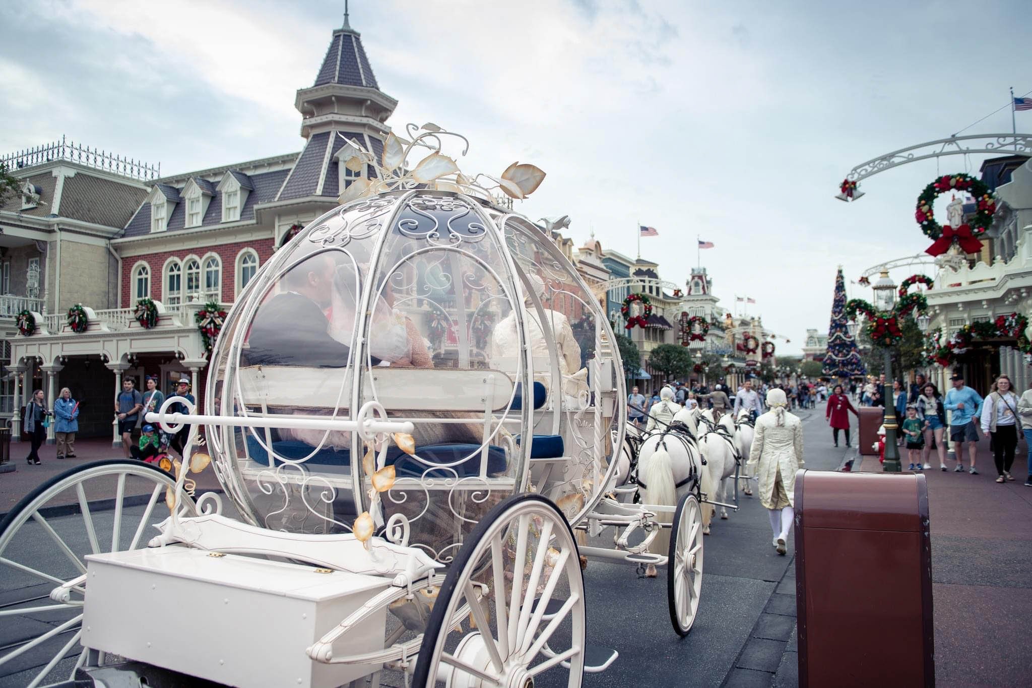 A white ornate horse-drawn carriage with a glass dome, decorated with a bow on top, stationed on Main Street at Disney's Magic Kingdrom decorated with wreaths and flags. People are walking and shopping in the background on a cloudy day Disney World.