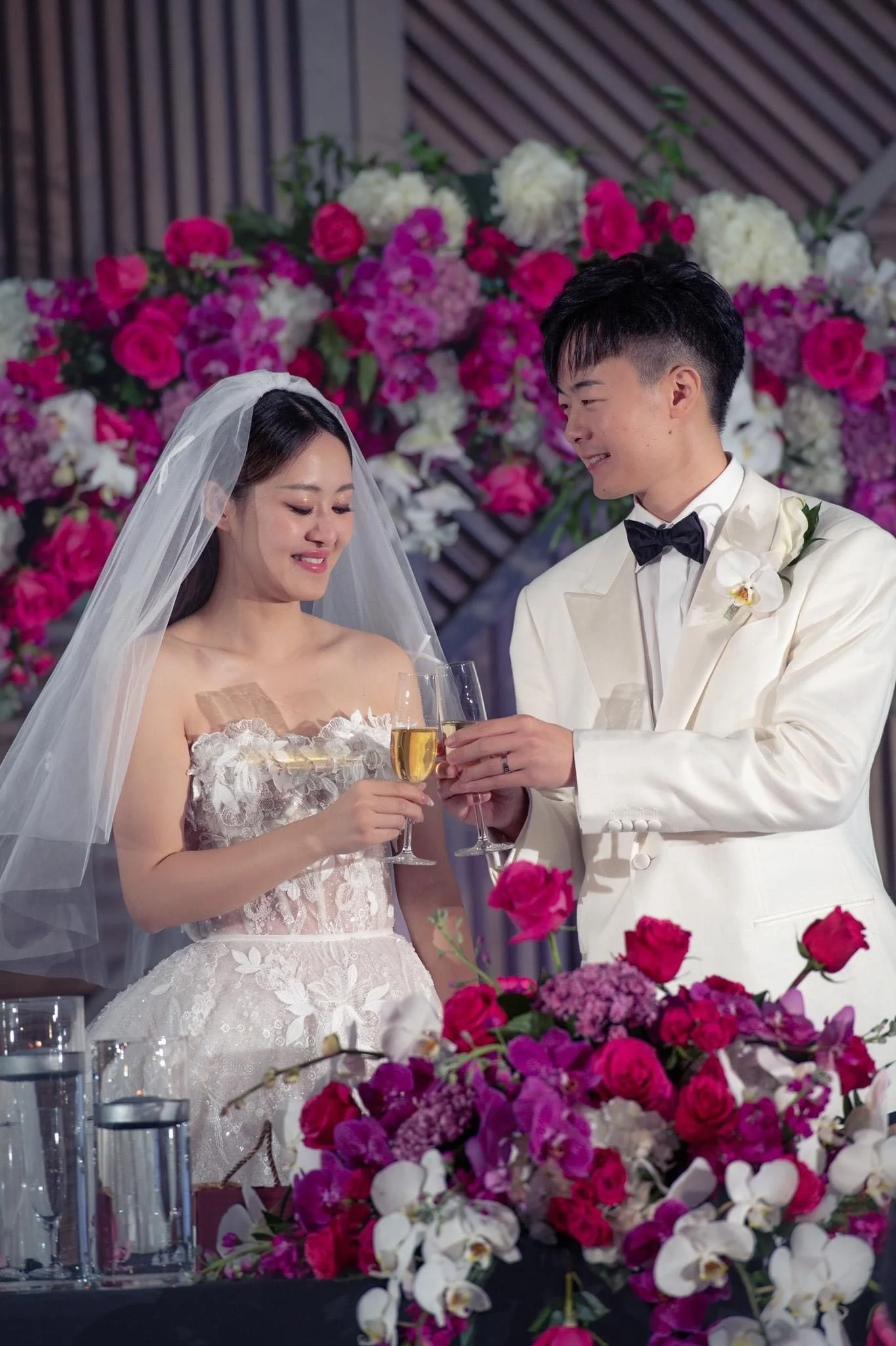 A bride and groom at their wedding, holding champagne glasses and smiling, with a backdrop of colorful flowers.