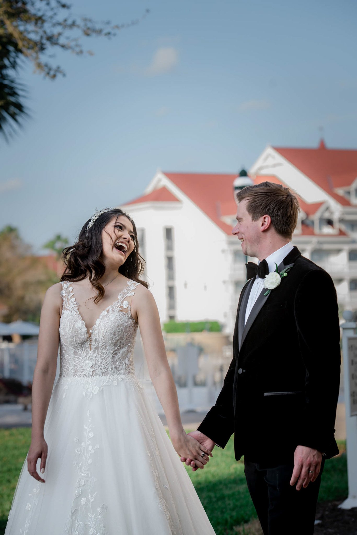 A bride and groom holding hands and smiling at each other outside on a sunny day, with a large white house with red roofs in the background.