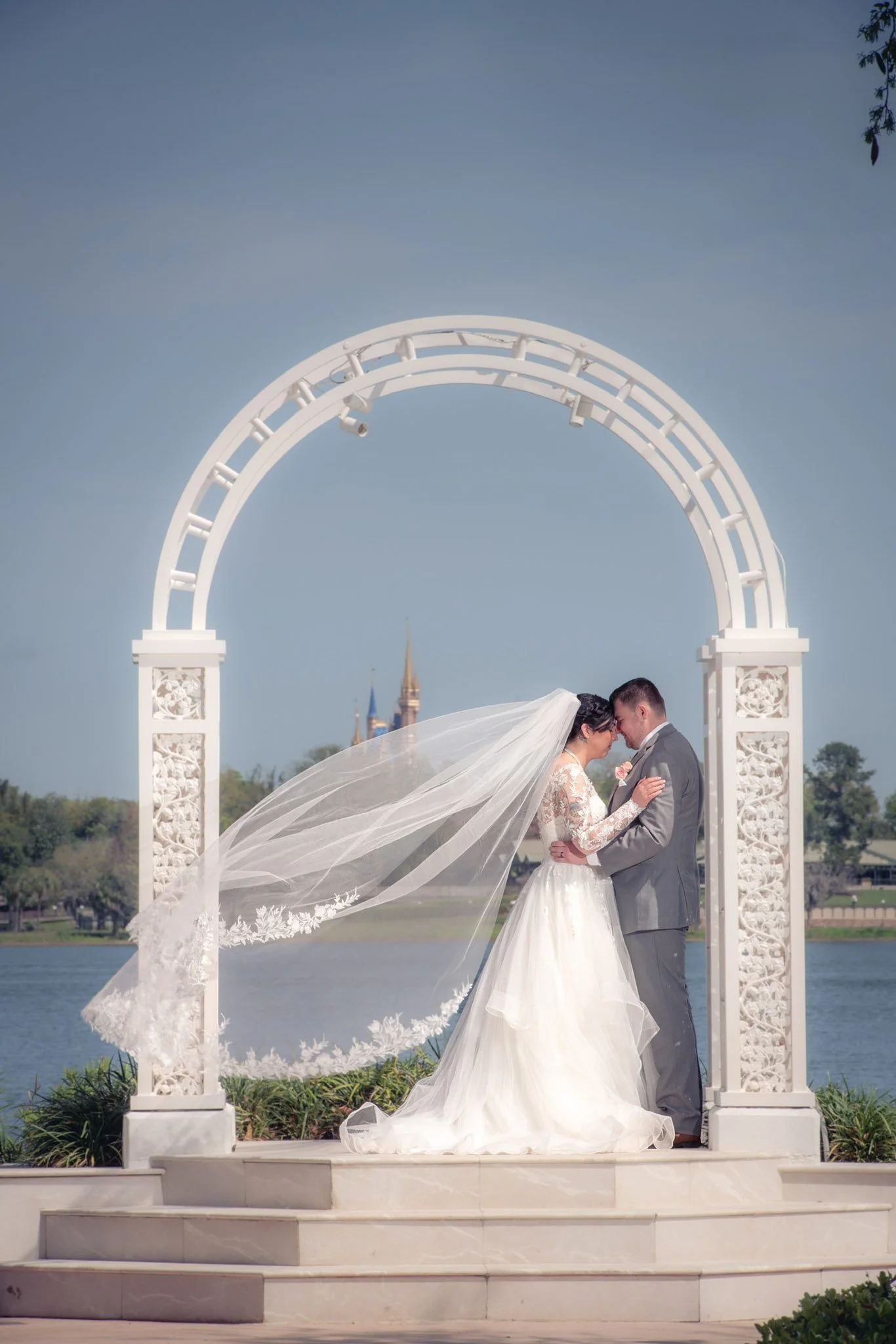A bride and groom on their wedding day under a white arch at Picture Point of Disney's Wedding Pavilion near a body of water with a castle in the background, the bride's veil flowing in the wind.