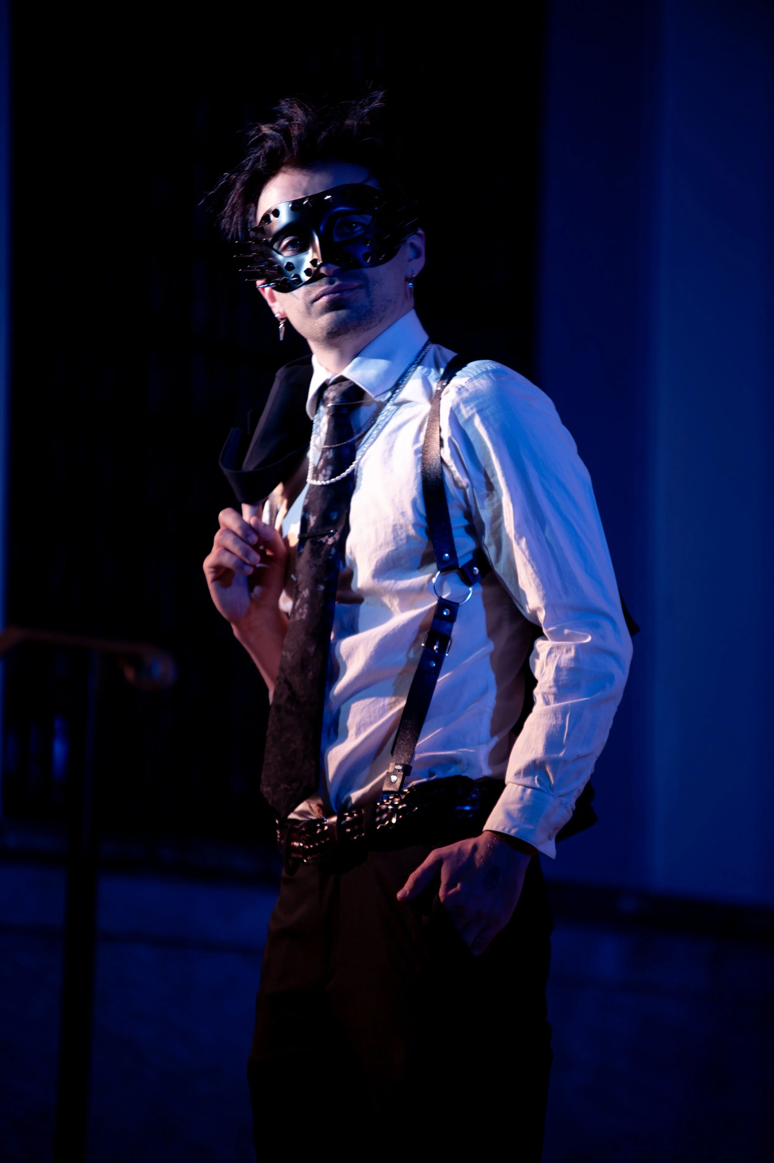 A person wearing a black mask, white shirt, black tie, suspenders, and black pants stands on a dimly lit stage with a dark background.