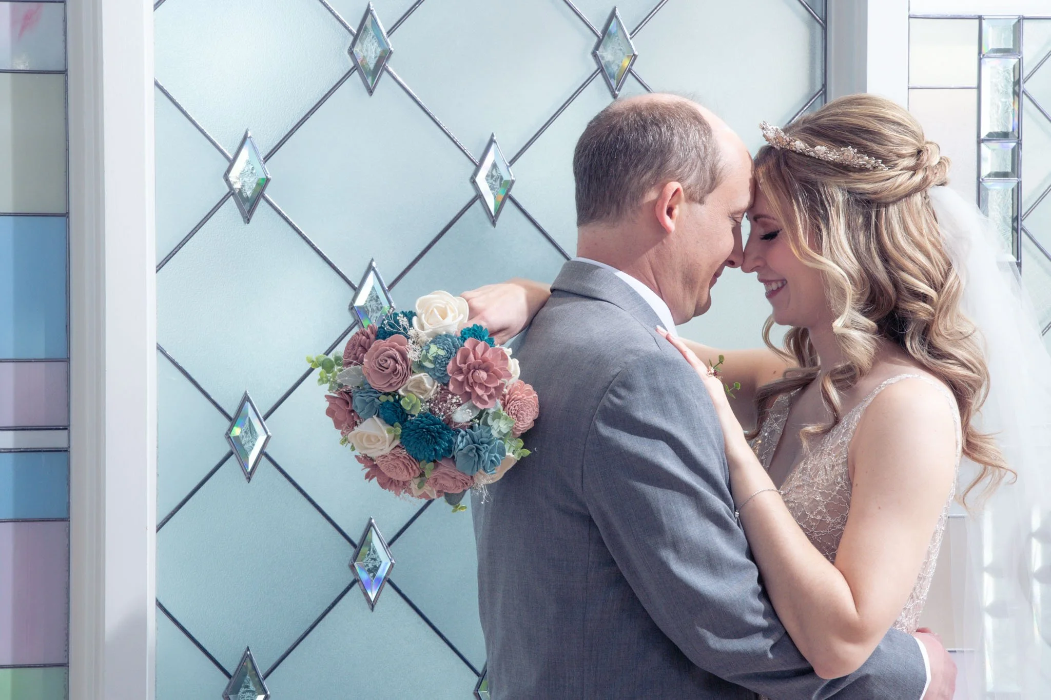A wedding couple, a man in a gray suit and a woman in a lace wedding dress, embracing in front of stained glass windows. The woman is holding a bouquet of pastel-colored flowers.