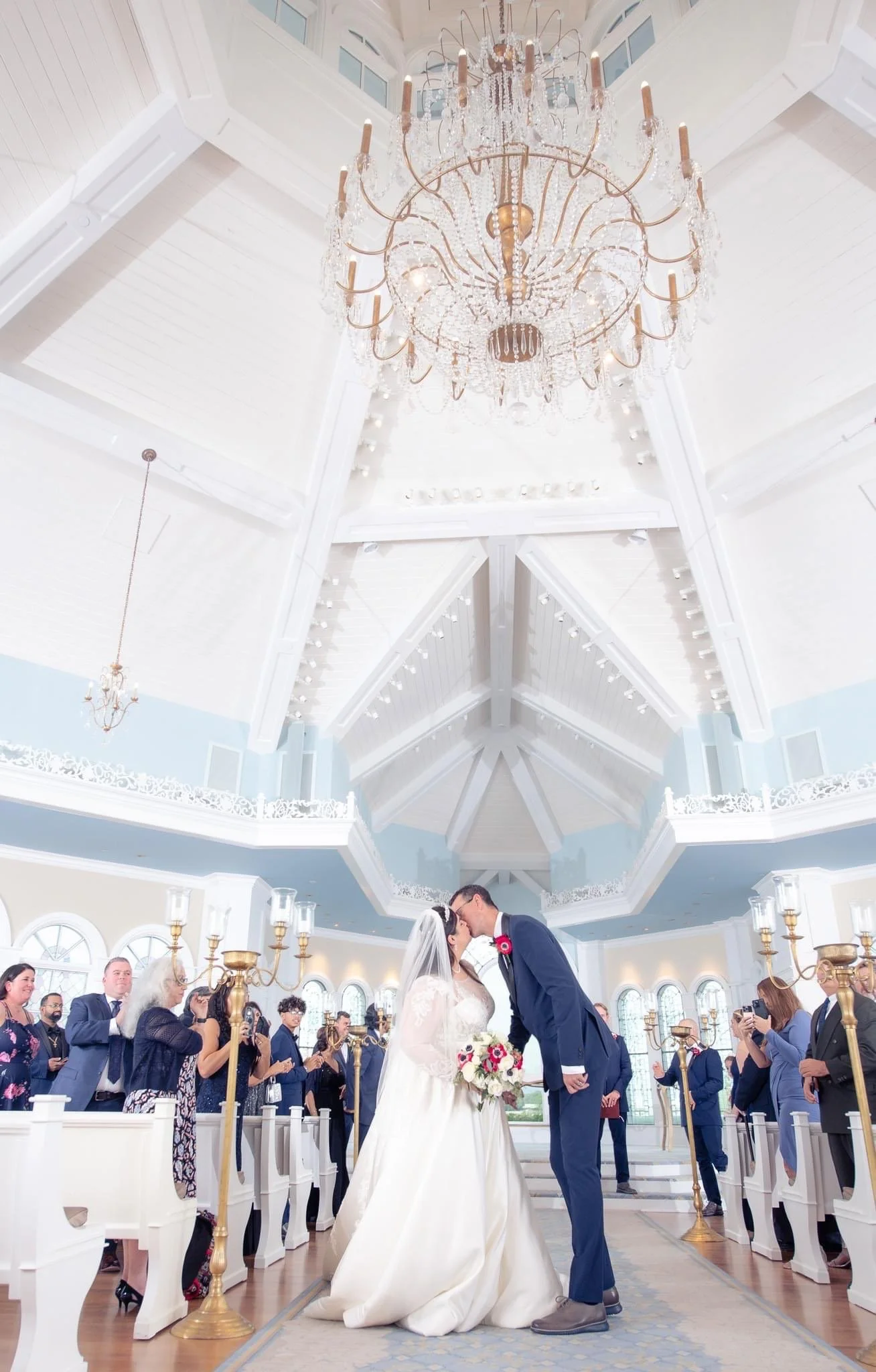 A bride and groom kiss during their wedding ceremony in Disney's Wedding Chapel with a large chandelier overhead and guests seated on either side.