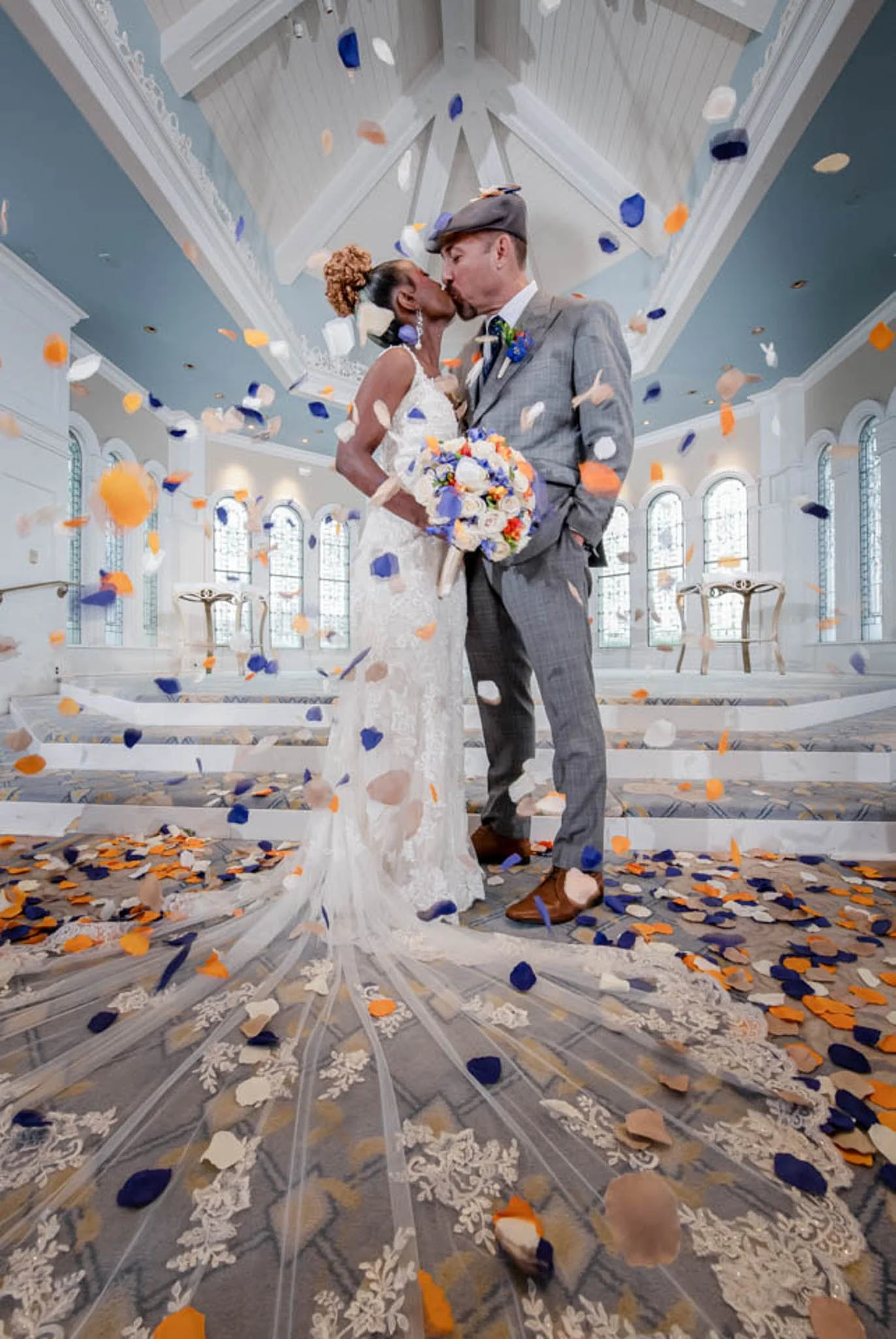 A bride and groom sharing a kiss, standing on a stage surrounded by falling confetti in an elegant, well-lit church or hall. The bride holds a bouquet, and both are dressed in wedding attire at Disney's Wedding Pavilion.