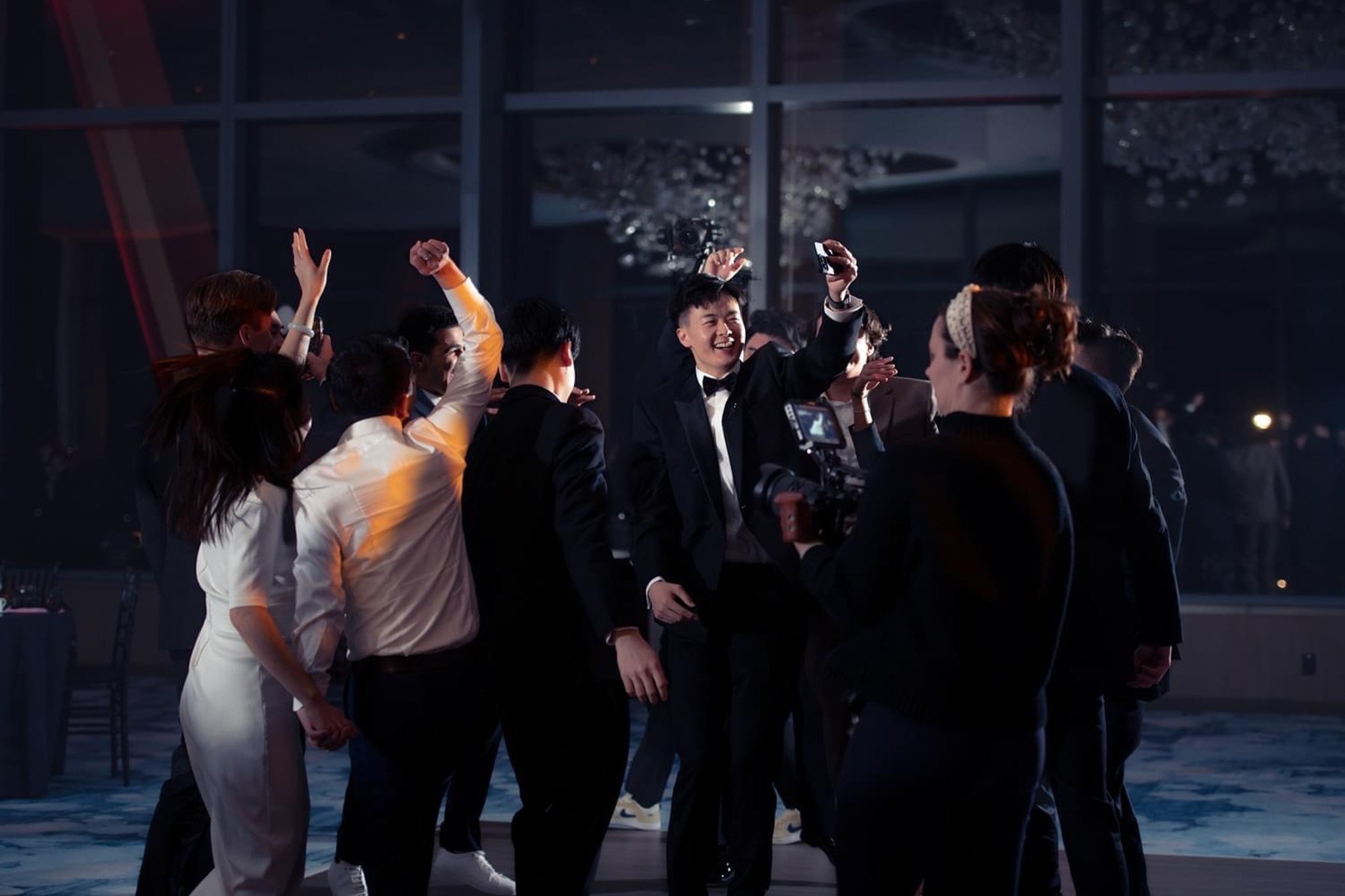 People dancing and celebrating at a formal event, with a man in a tuxedo taking a selfie with a group, in a large indoor venue with glass windows.
