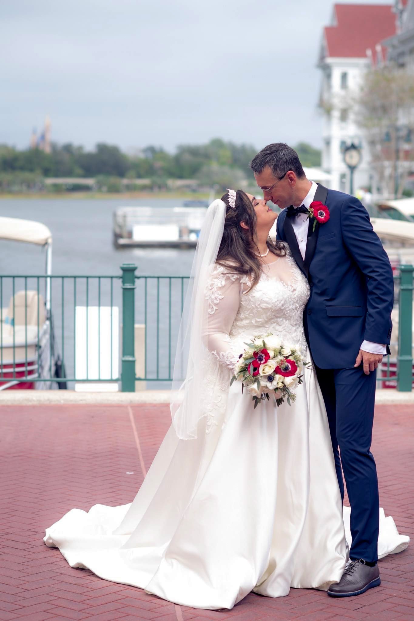 A bride and groom sharing a kiss near a waterfront, with boats and The Grand Floridian in the background.
