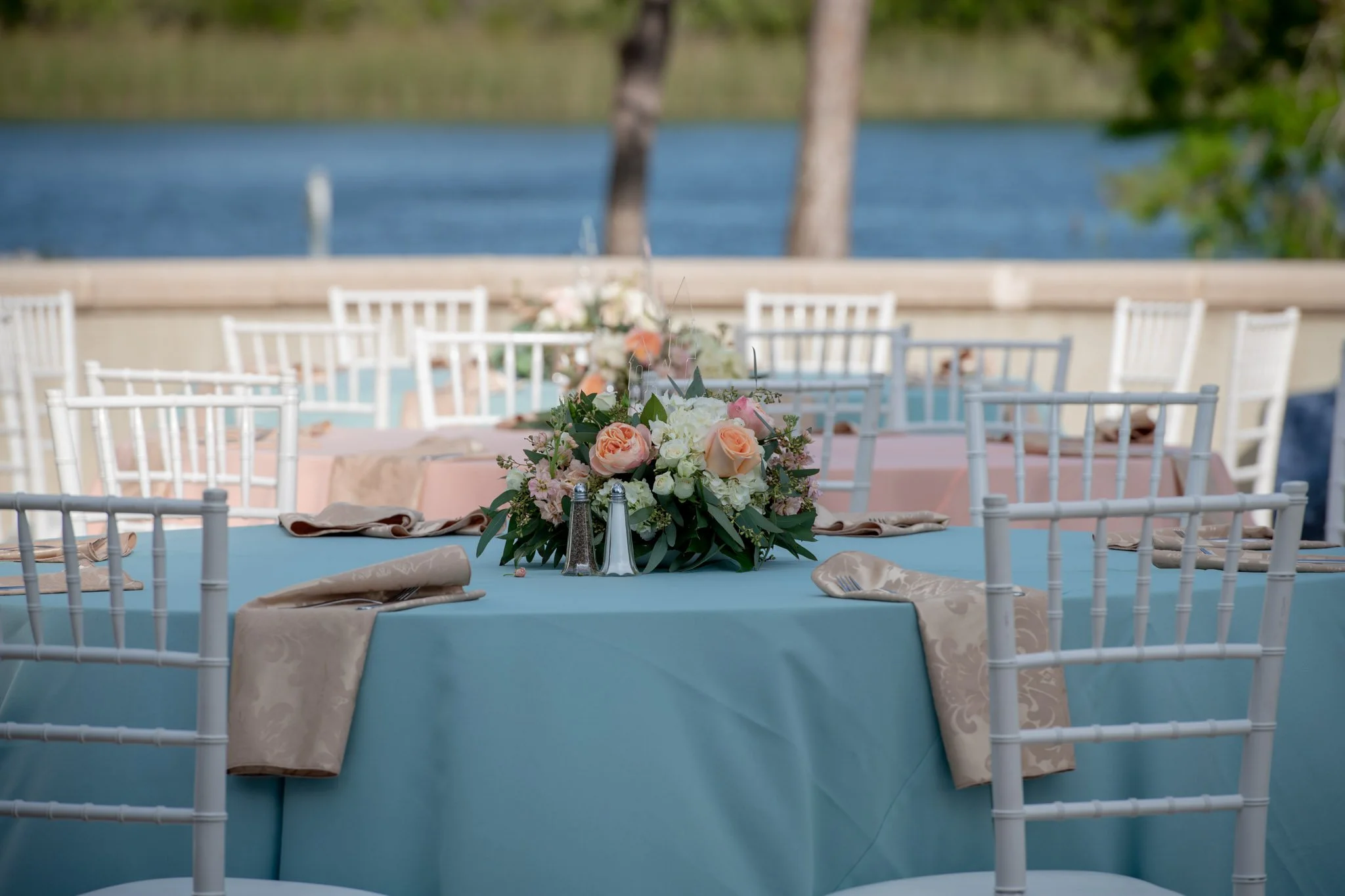 Elegant outdoor event table with pastel-colored tablecloths, floral centerpiece, and white chairs on a terrace overlooking a body of water and trees.