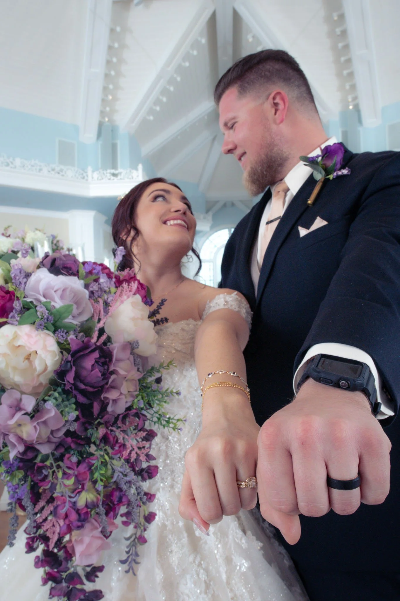 A bride and groom during their wedding, holding hands and smiling at each other. The bride is holding a large bouquet of purple, pink, and white flowers. The groom is dressed in a dark suit with a boutonnière, and they are in a bright, elegant venue 